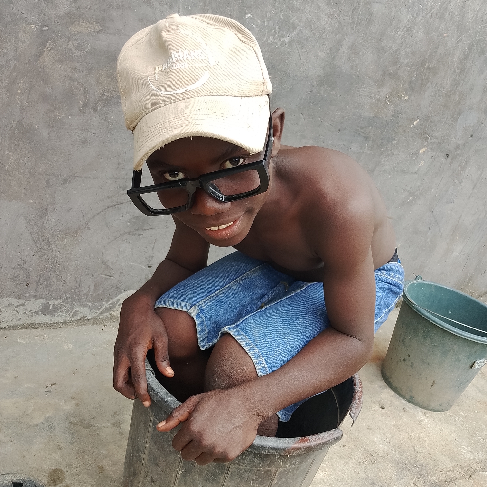 A young, shirtless boy wearing a light-colored baseball cap and oversized black-rimmed glasses squats inside a large, dark-colored bucket. He is also wearing blue denim shorts. His hands are resting on the rim of the bucket. A second, lighter green bucket sits to the boy's right. The background is a textured, gray concrete wall, and the ground is also concrete. The scene appears to be outdoors, possibly in a yard or an open area in Ado, Nigeria.