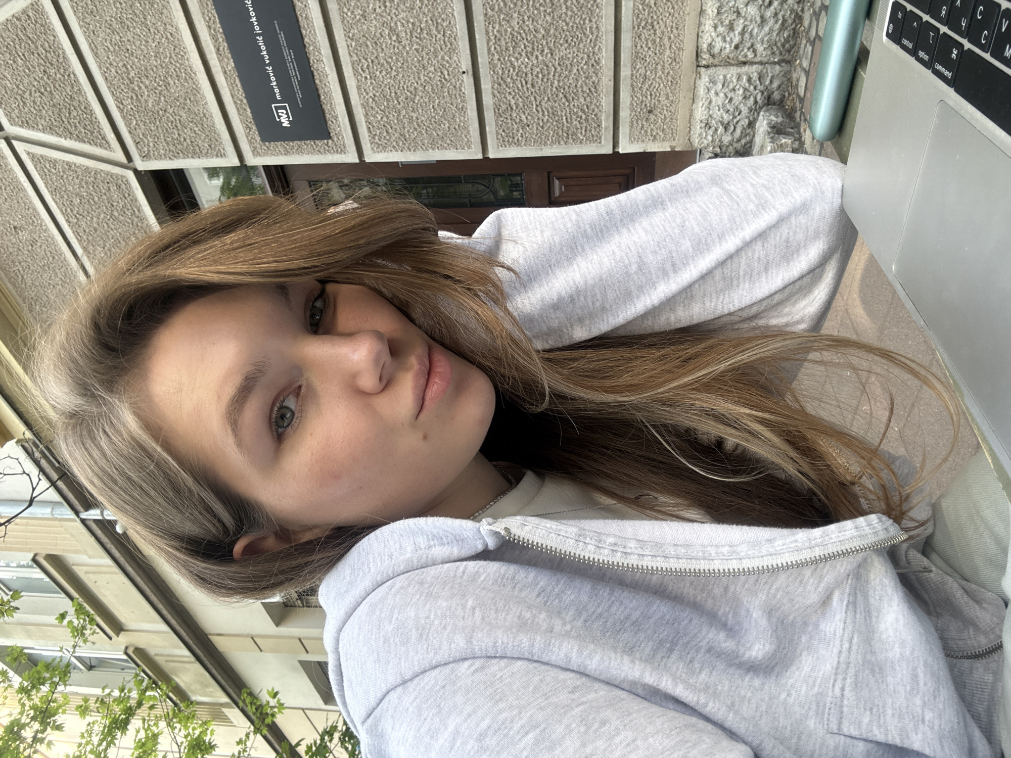 A young woman is positioned outdoors in Belgrade, Serbia. She is fair-skinned with light brown hair. She is wearing a light gray zip-up hoodie over a white shirt, and a thin silver necklace. She is sitting at a table and using a laptop. The building behind her is beige and brown with a sign that says "markovic vukovic jovkovic" in Serbian.