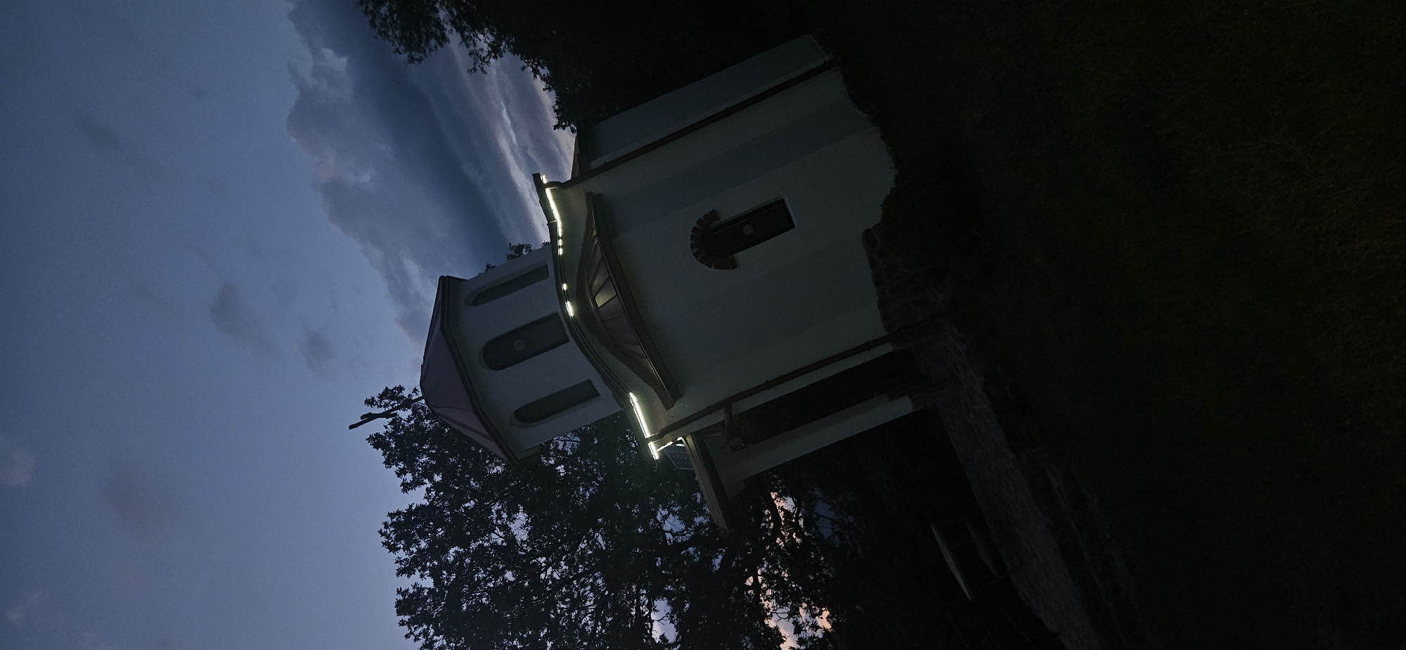 A small, white-walled Orthodox church stands at dusk or early night in Gutsal, Bulgaria, captured from a low, tilted perspective. The structure features a multi-tiered, reddish-brown tiled roof with a central octagonal bell tower surmounted by a cross. Three arched windows are visible on the bell tower, and a single arched window with brick detailing is present on the main facade. Warm-white LED strip lighting is installed along the guttering beneath the roof eaves, illuminating the upper portion of the church. A small solar panel is visible on the roof section above the illuminated guttering. Dense, dark foliage from trees surrounds the church, particularly on the left. The sky is a deep indigo blue with visible clouds, showing a faint warm glow on the far left horizon. The ground in the immediate foreground is dark and indistinct, likely grass, with a stone foundation visible at the church's base.