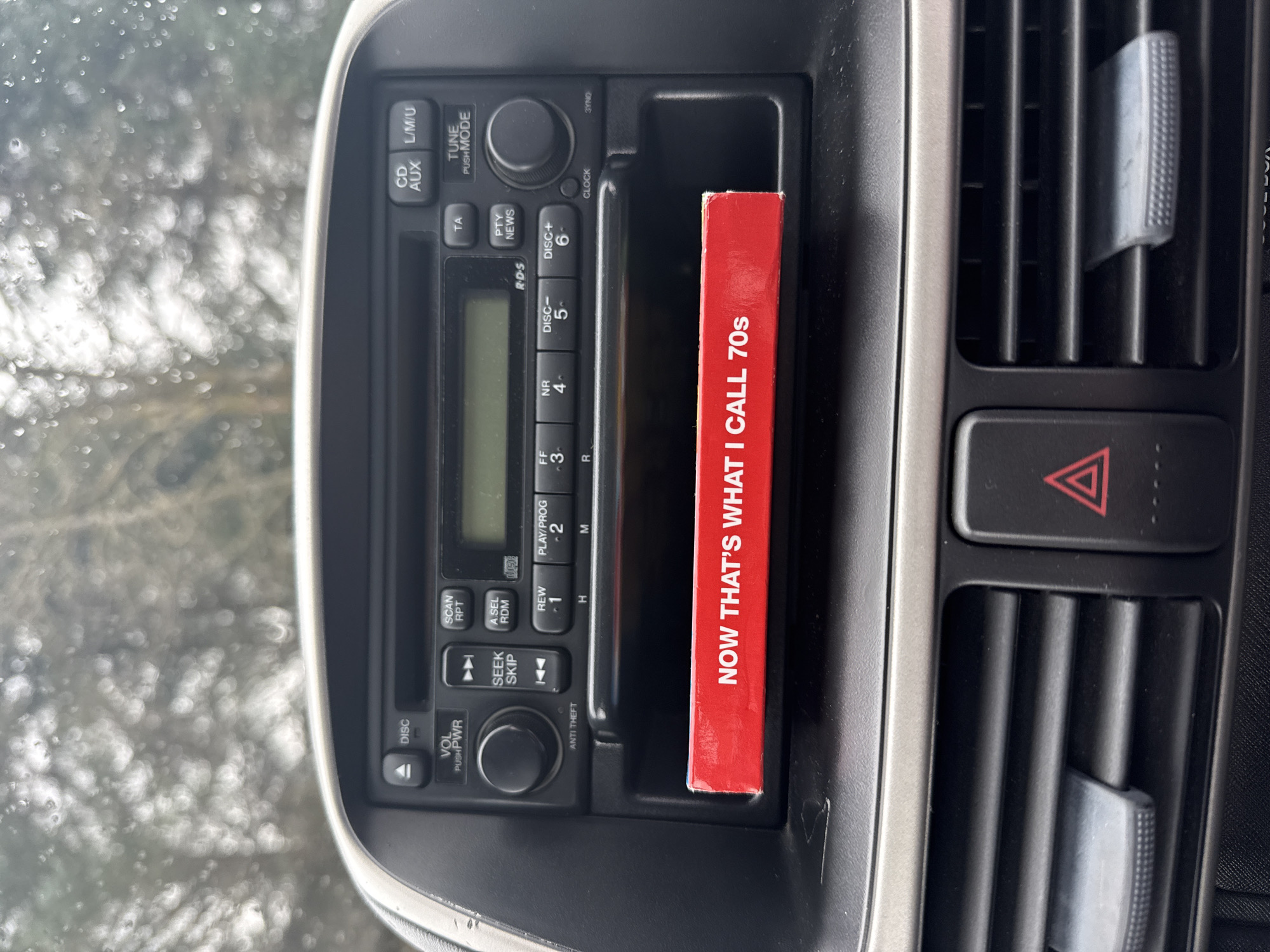 The image shows the center console of a vehicle, specifically focusing on the radio and adjacent vents. The radio is a standard car stereo with buttons for CD, AUX, tuning, scanning, seeking, and volume/power. A red CD case labeled "NOW THAT'S WHAT I CALL 70s" is inserted into a slot or compartment next to the radio. To the right of the radio are air vents with adjustable louvers, and below them is a hazard light button. The location in the city of Unknown, Unknown is unknown.