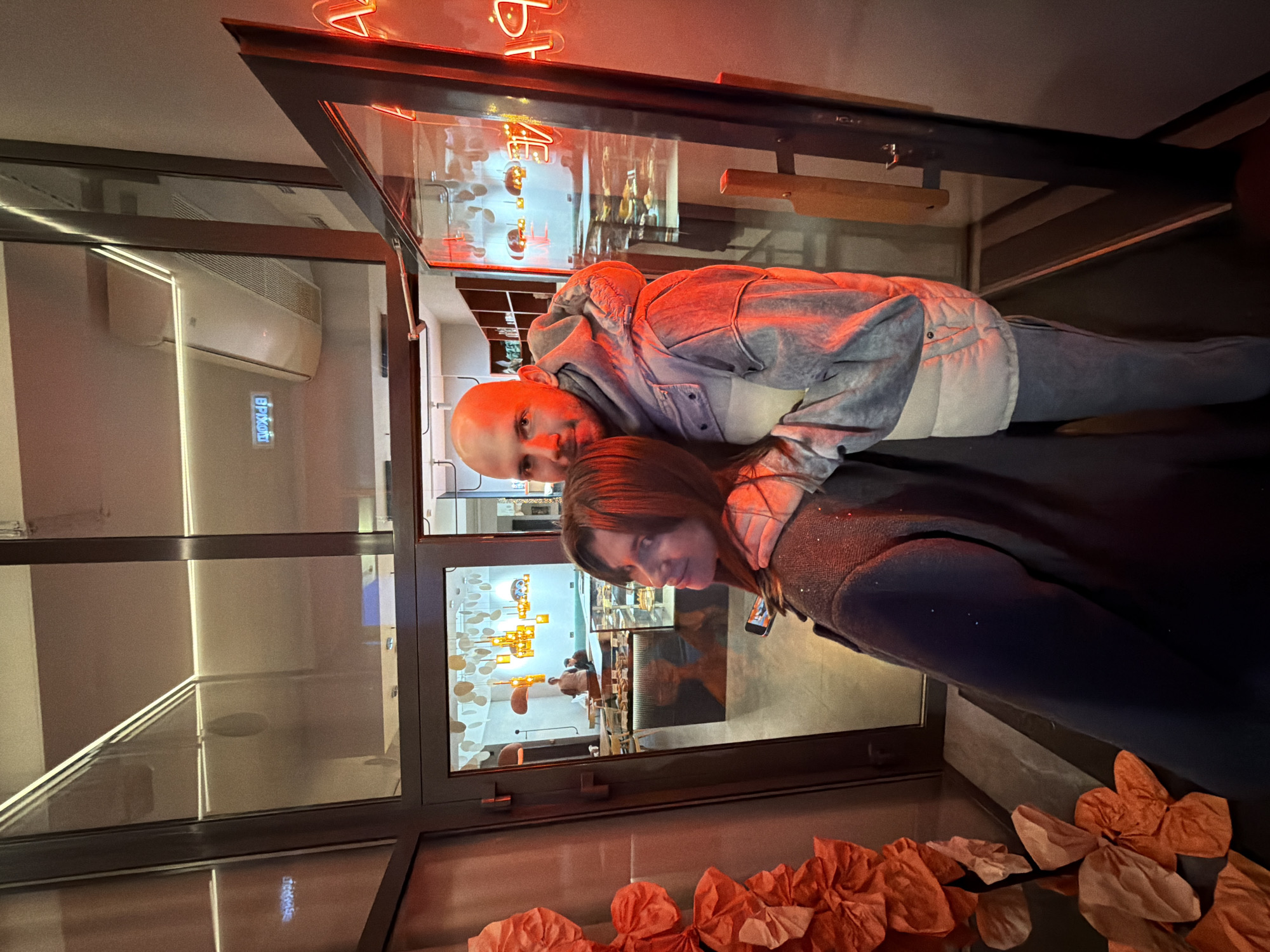 A bald man in a gray and white jacket and a woman in a black jacket are posed in front of a large window with a black frame, decorated with orange paper flowers at the bottom. Reflected in the window is the interior of a brightly lit building with a glass display case and chandelier-style lighting. The man has his arm around the woman's neck. The image appears to be taken at a slight angle. This location is in Krasnoyarsk, Russia.