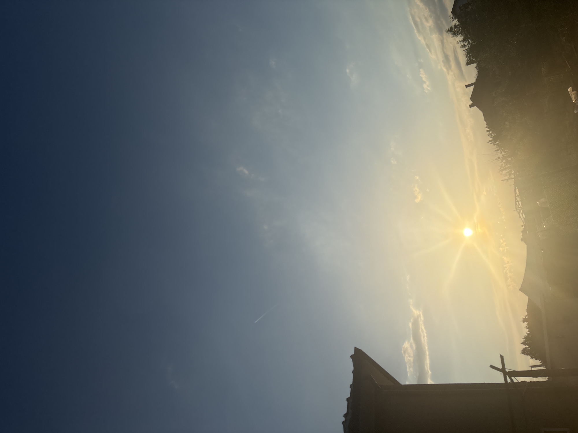 The image depicts a view of the sky with the sun shining brightly. Silhouetted structures, possibly buildings and vegetation, are visible in the lower portion and along the right side. A faint contrail from an aircraft is present in the sky. The sky transitions from a deep blue on the left to a lighter, sunlit hue on the right.