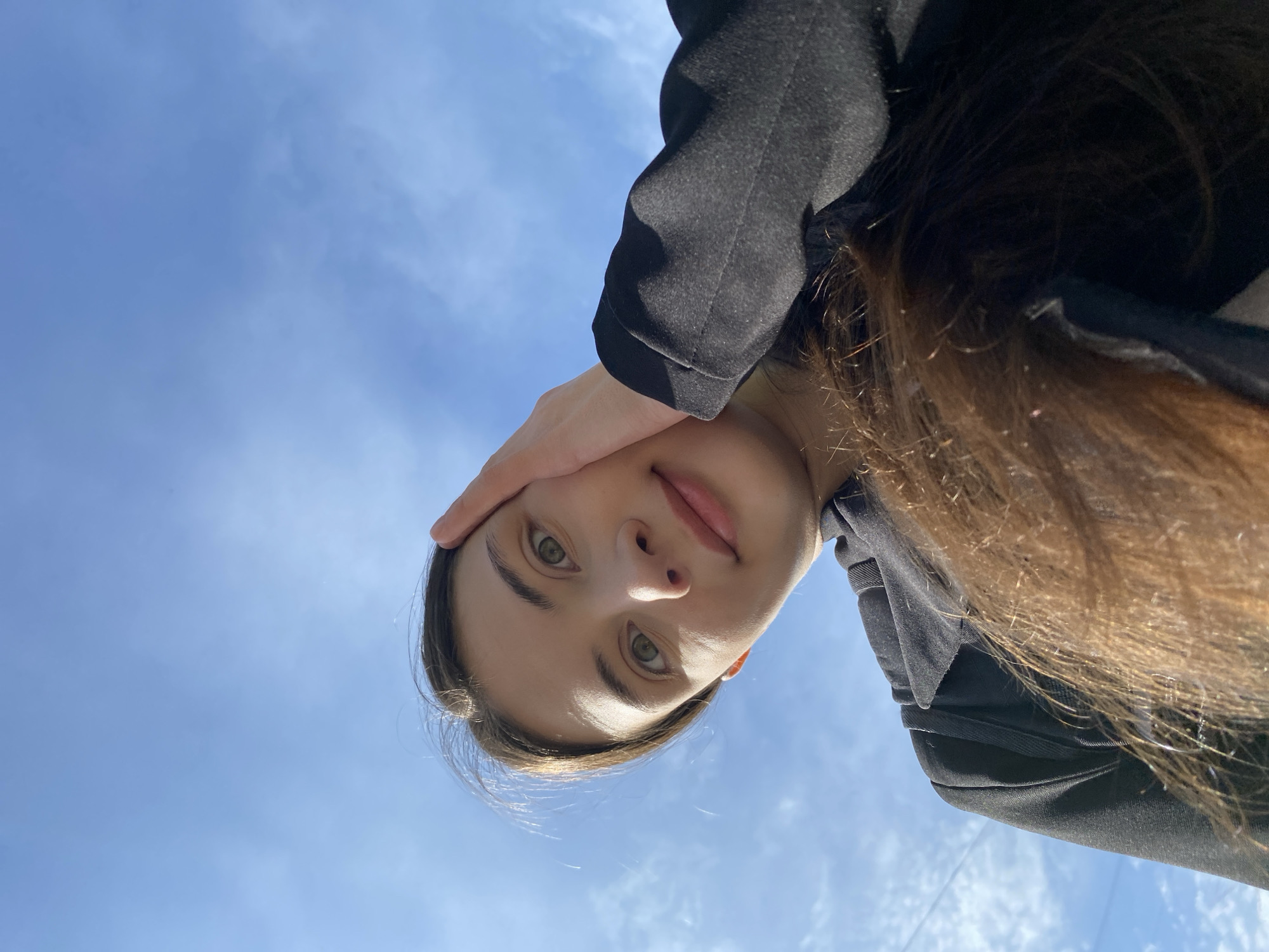 A woman with light skin, brown hair, and green eyes is looking down at the camera from a low angle. Her right hand is touching her forehead. She is wearing a black jacket. The background is a bright blue sky with some white clouds. The photograph appears to have been taken outside during daylight hours, but there is insufficient information to state with confidence the exact location in Votkinsk, Russia.