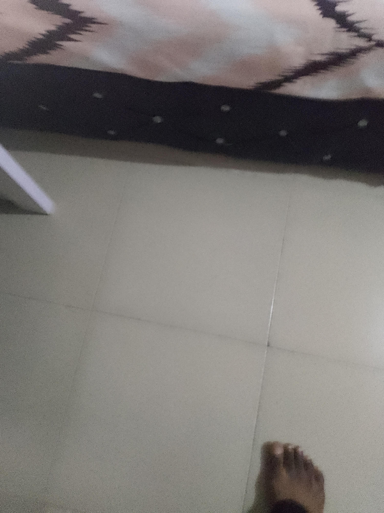 The image shows a person's foot on a tiled floor. The floor appears to be light gray with visible grout lines. In the background, a dark-colored object with a patterned texture is partially visible. The overall lighting is dim. The scene is not specific to any particular location within the city of Jauro Yinu, Nigeria, as there are no identifiable landmarks or contextual elements.