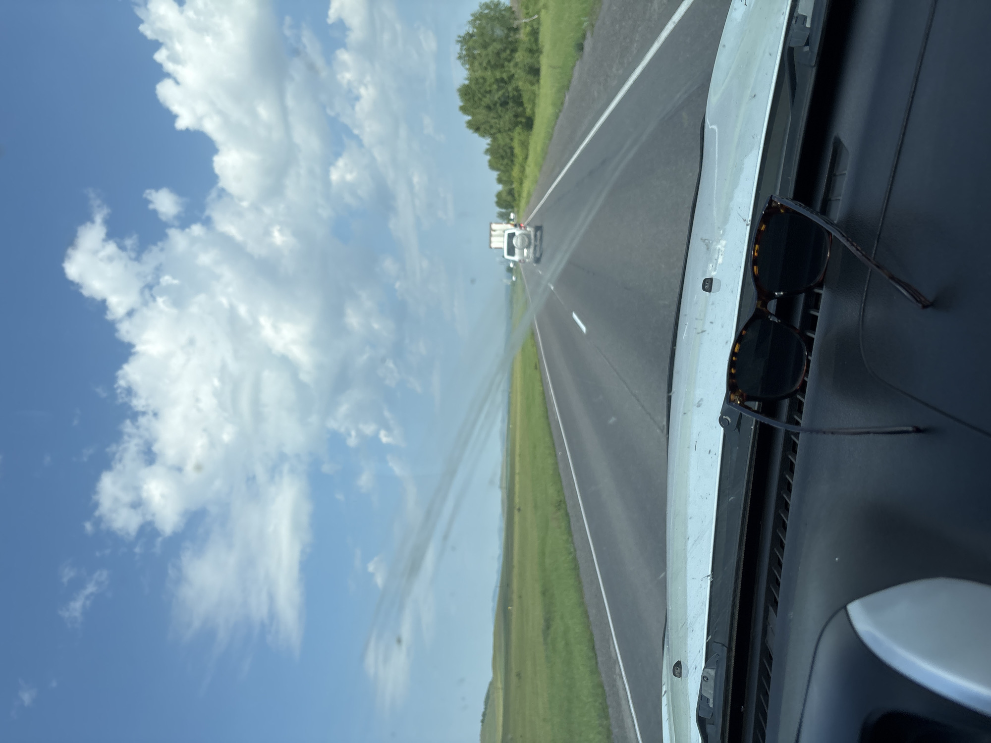 The image depicts a view from inside a vehicle looking out at a highway. The foreground shows a car's dashboard with a pair of sunglasses resting on it. The road stretches into the distance, with other vehicles, including a white SUV and a truck, visible on it. The landscape on either side of the road consists of green fields and trees under a blue sky with white clouds. The scene suggests a journey on a sunny day in an open, rural area. Based on the image alone, the specific location in Borodino, Russia cannot be determined.