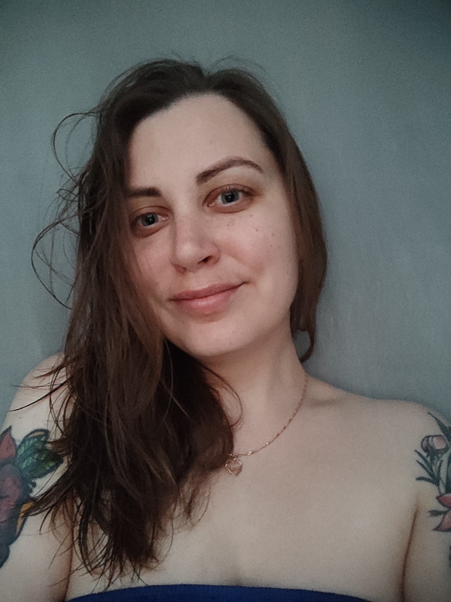 A woman with brown hair and blue eyes is shown from the chest up, with a grey wall in the background. She is wearing a gold chain with a heart-shaped pendant and has tattoos on her shoulders. Her hair is slightly messy and falling around her face. She is looking directly at the camera with a slight smile. The lighting is soft and even.