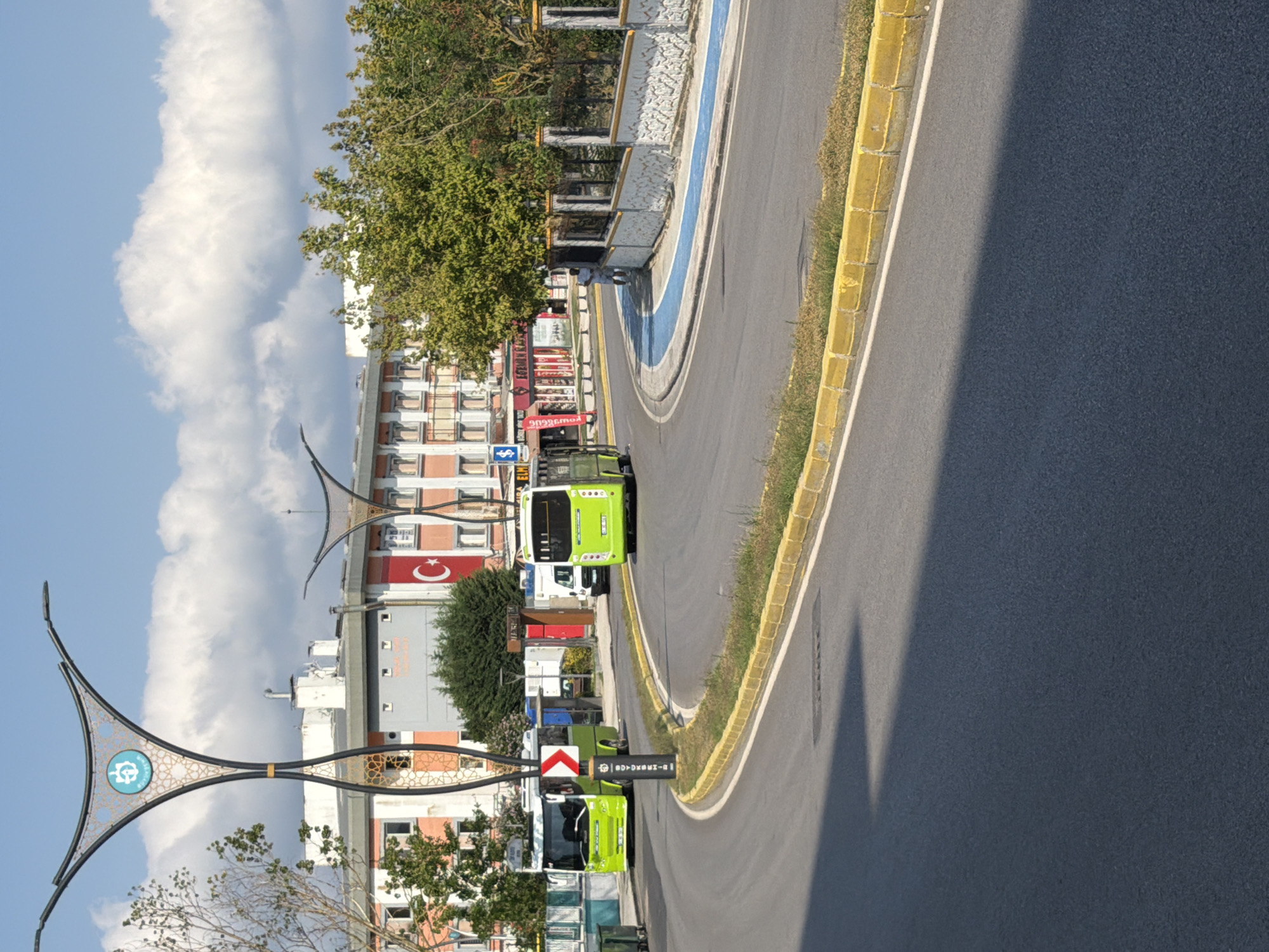 The image depicts a street scene in the city of Kocaeli, Turkey. In the foreground, a paved road curves to the right, bordered by yellow curbs and some vegetation. In the middle ground, two bright green buses are visible on the road. The bus on the left is turning, indicated by a directional sign. The bus on the right is further down the road. Behind the buses, there are buildings with a Turkish flag displayed on one of them. Decorative streetlights with intricate patterns are prominent on the left side of the image. A pedestrian pathway with a blue surface runs alongside a stone wall on the right. The sky is blue with scattered white clouds. The overall scene suggests a public transportation hub or a busy street in an urban area.
