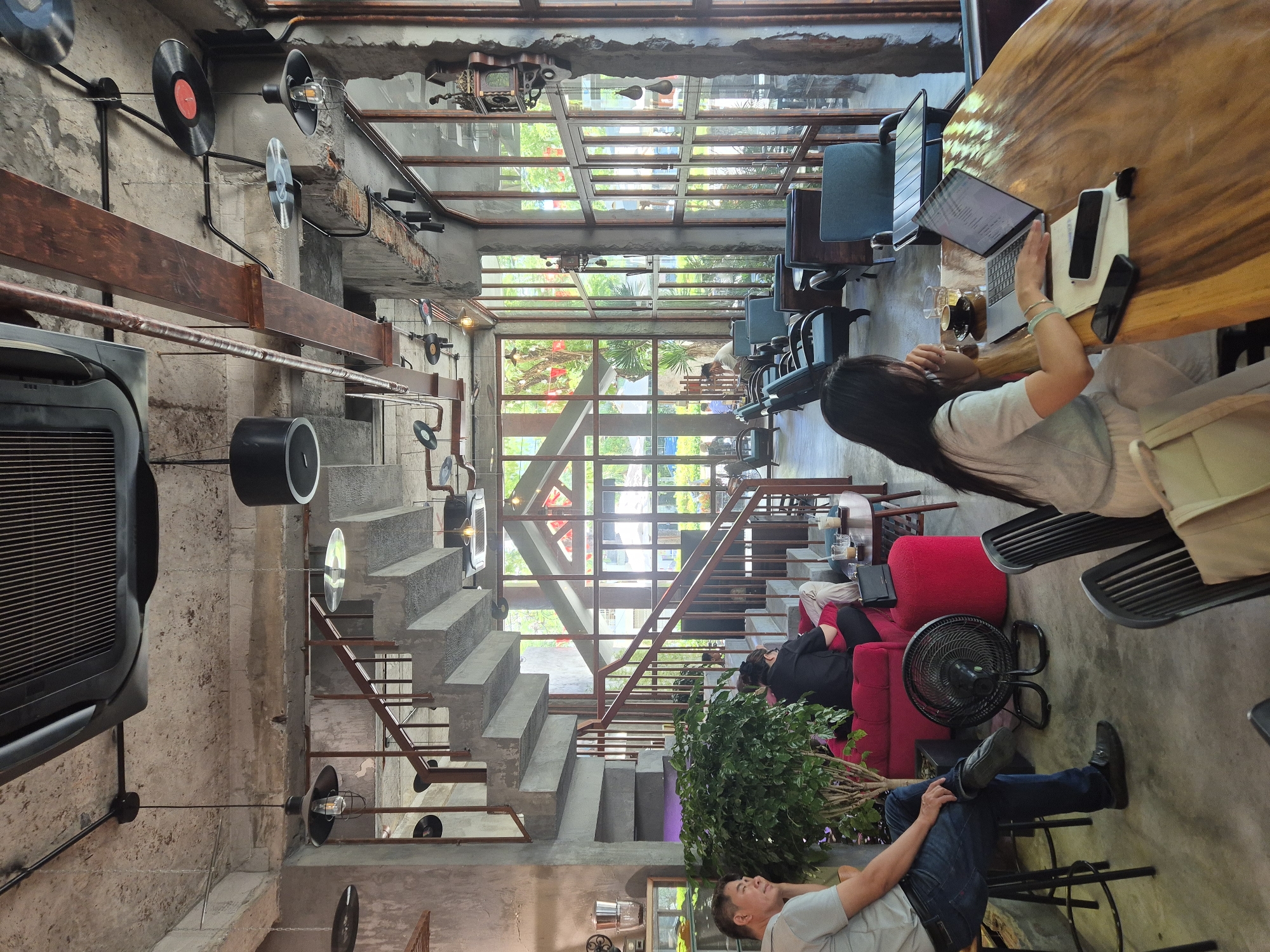 This image depicts the interior of a contemporary cafe located in Đà Nẵng, Vietnam, characterized by an industrial-chic aesthetic. Exposed concrete walls and ceilings, along with rough-hewn wooden beams and metal conduits, create a raw and unfinished ambiance. A prominent feature is a sweeping concrete staircase with metal railings, leading to an upper level.
Several patrons are visible throughout the space. In the foreground, a man in a grey t-shirt and jeans is seated on a stool, his legs crossed, looking towards the right. To his left, a vibrant red sofa is occupied by a person in black. Near the sofa, a black oscillating fan stands on the concrete floor.
Further into the cafe, a woman in a light-colored shirt is engrossed in her laptop at a large, polished wooden table. A coffee cup and a glass of water are beside her. On the table, two smartphones and a notebook are also present. Other seating areas include plush blue armchairs arranged near large, multi-paned windows that offer views of lush greenery outside.
The lighting is a mix of industrial pendant lamps with exposed bulbs and recessed ceiling lights, contributing to the cafe's moody and atmospheric setting. Decorative elements include vintage vinyl records suspended from the ceiling, adding a retro touch to the modern design. The overall scene suggests a relaxed and productive environment, popular with individuals seeking a comfortable space for work or leisure.