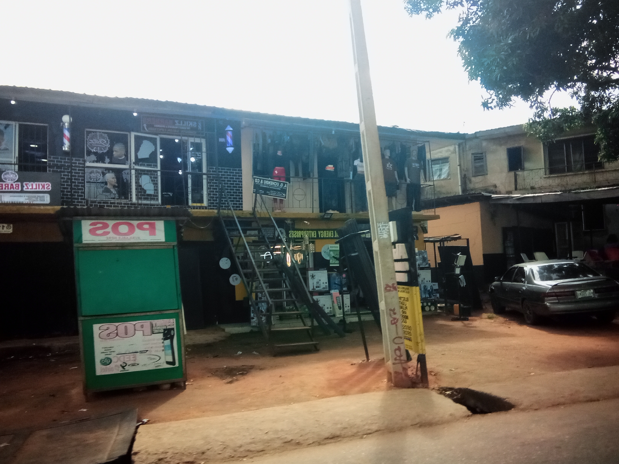 In Nibo, Nigeria, a street scene unfolds in front of a two-story building with several businesses. The ground floor features a barber shop with a barber pole outside, mannequins visible through the window, and various posters. Adjacent to the barber shop is a green POS stand with advertisements. Further down, an external metal staircase leads to the second floor, where more storefronts and balconies are present, with mannequins displayed on the upper level. A dark-colored sedan is parked on the dirt road in front of the building. A utility pole with advertisements stands prominently in the foreground. The sky is overcast, and the ground is a dusty, reddish-brown earth.