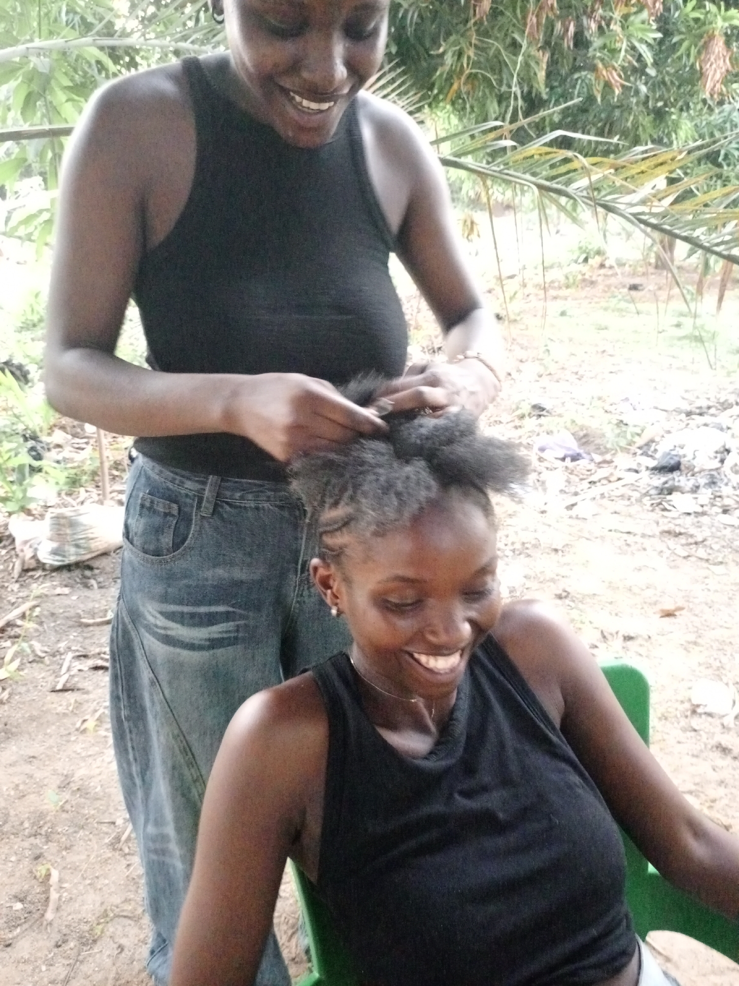 Two dark-skinned women are outdoors in Keffi, Nigeria. One stands behind the other, who is seated in a green plastic chair. The standing woman is braiding the seated woman's hair. Both women are smiling and wearing black tank tops. The standing woman is also wearing jeans. The ground is dirt and there is sparse vegetation visible in the background.