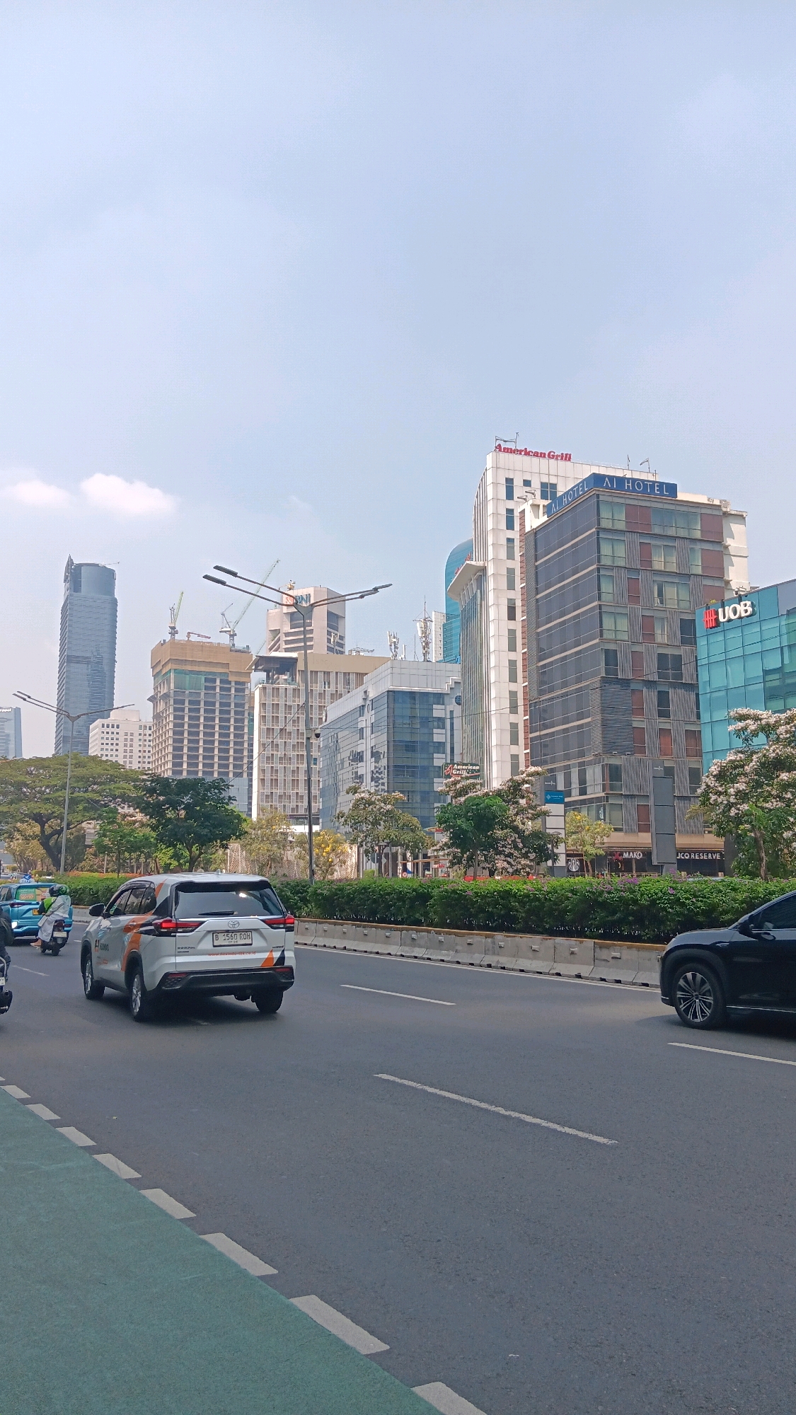 The image shows a view of a city street in Central Jakarta, Indonesia, with modern high-rise buildings and moderate traffic. In the foreground, a white SUV with an orange stripe is driving on the right side of the road, alongside a green bicycle lane. Several other vehicles, including motorcycles and cars, are visible on the road. Behind the vehicles, a row of lush green bushes separates the road from a sidewalk lined with trees, some of which are in bloom with white flowers.
The background is dominated by a cluster of contemporary skyscrapers, varying in height and architectural style. One prominent building features a red sign that reads "American Grill" and "AI HOTEL" in blue letters. Another building displays the "UOB" logo. Construction cranes are visible behind some of the buildings, suggesting ongoing development. The sky is a light blue with a few scattered clouds. The overall impression is of a bustling urban environment with a mix of business and residential structures.