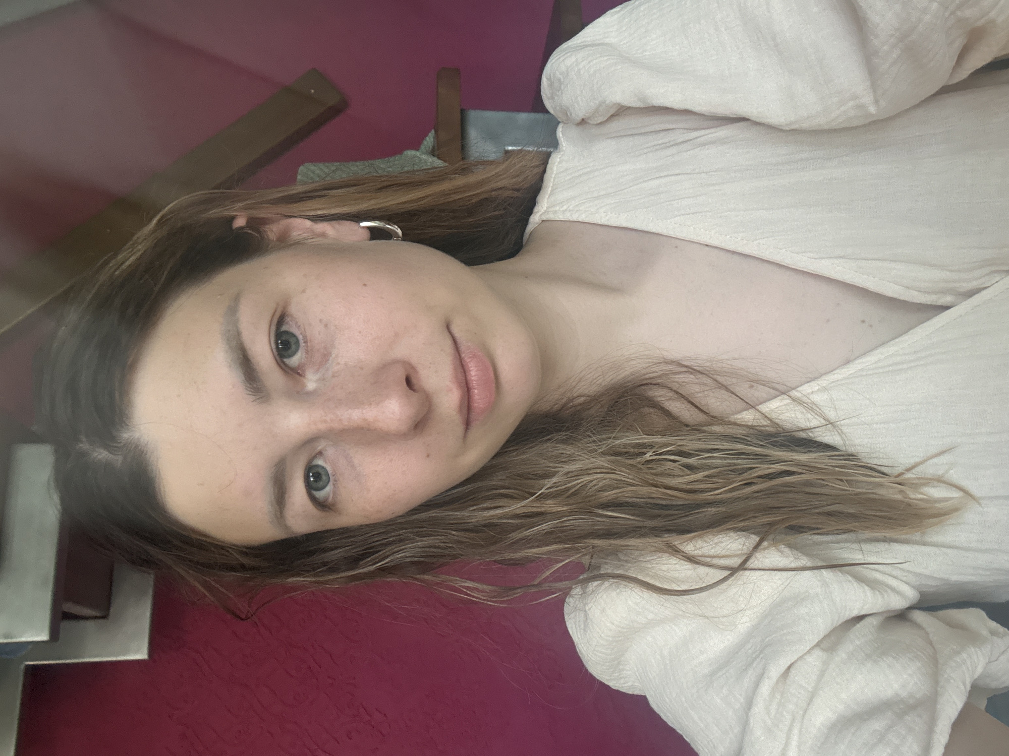 A woman with light skin, brown hair, and gray eyes is positioned at an oblique angle in the image. Her face features visible freckles, particularly on her nose and cheeks. She is wearing a light-colored shirt with a textured pattern. A silver hoop earring is visible in her left ear. The background consists of a red wall, a wooden object, and what appears to be a stainless steel structure. There is no indication that the photo was taken in the city of Чёрное, Russia.