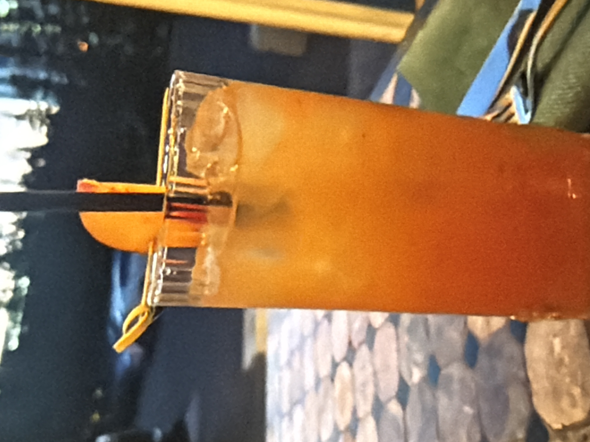 The media file shows a close-up, horizontally oriented shot of a drink in a tall glass. The drink is a vibrant orange color and appears to be a cocktail or juice, possibly with ice. A black straw is visible inserted into the drink, along with a slice of orange or lemon as a garnish, attached with a thin yellow loop. The glass itself has a textured rim.
The background is dark and somewhat blurred, suggesting an indoor setting, possibly a bar or restaurant. Hints of a patterned surface, possibly a tablecloth or placemat, are visible at the bottom of the frame, with a circular, mosaic-like design in muted blues and grays. A portion of a green cloth or garment is also visible in the upper right corner, with a blue and white striped ribbon or fabric draped over it.
The lighting suggests it might be evening or indoors with artificial lighting. No people are clearly visible, and no text is discernible. The overall impression is a still life of a refreshing beverage.