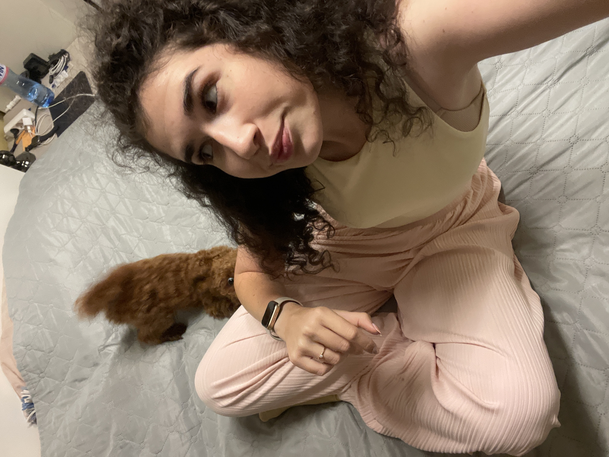 A person with dark, curly hair is sitting on a gray, quilted bed. They are wearing a light yellow top and light pink pleated pants. A small, brown, curly-haired dog is also on the bed, with its back to the camera, in the upper left portion of the frame. The person is looking off to the right of the frame with a slight smile. In the background, on a nightstand, there is a bottle of water, a phone charger, and other electronic accessories. The scene appears to be indoors, possibly in a bedroom in Sofia, Bulgaria.