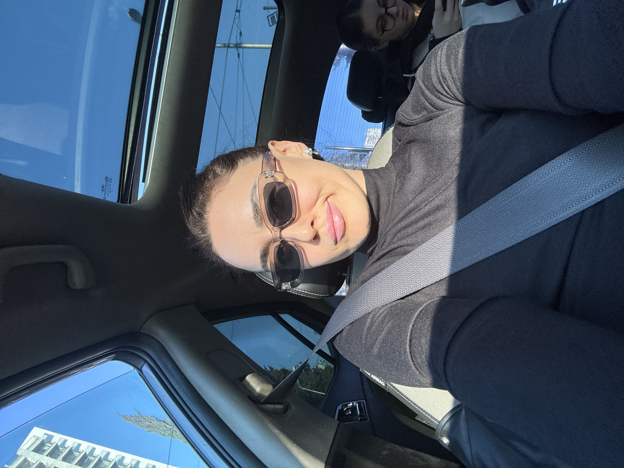The image depicts a selfie taken from the front passenger seat of a vehicle on a bright, sunny day in an urban environment of Tbilisi, Georgia.
In the foreground, a woman with dark hair pulled back is visible from the chest up, wearing clear-framed rectangular sunglasses with dark lenses, light pink lipstick, and a black turtleneck long-sleeve top. She has small, light-colored stud earrings and a slight smile. A grey seatbelt is diagonally across her chest and lap. Her right arm is subtly raised, indicating she is holding the camera.
In the back seat, partially obscured by the front passenger seat and the main subject's head, another individual, likely female, with dark hair and glasses, is visible. She appears to be looking down at a mobile phone held in her hands.
The vehicle interior features dark-colored upholstery and a panoramic glass sunroof extending overhead, revealing a clear blue sky. A light grey fabric insert is visible on the front passenger seat. Through the left side window, a tall, light-colored modern multi-story building with repetitive balcony structures is seen against the blue sky, along with a slender, dark green tree. Through the rear window and sunroof, more blue sky is visible, along with utility poles and wires typical of urban infrastructure. Strong sunlight casts distinct shadows within the car, particularly on the main subject's body and lap, emphasizing the bright external conditions.
The visible architecture and urban elements are consistent with developed residential or commercial areas within Tbilisi, such as those found in districts like Saburtalo or Vake, which feature modern apartment complexes and associated infrastructure. A precise street or neighborhood cannot be determined from the available visual information.