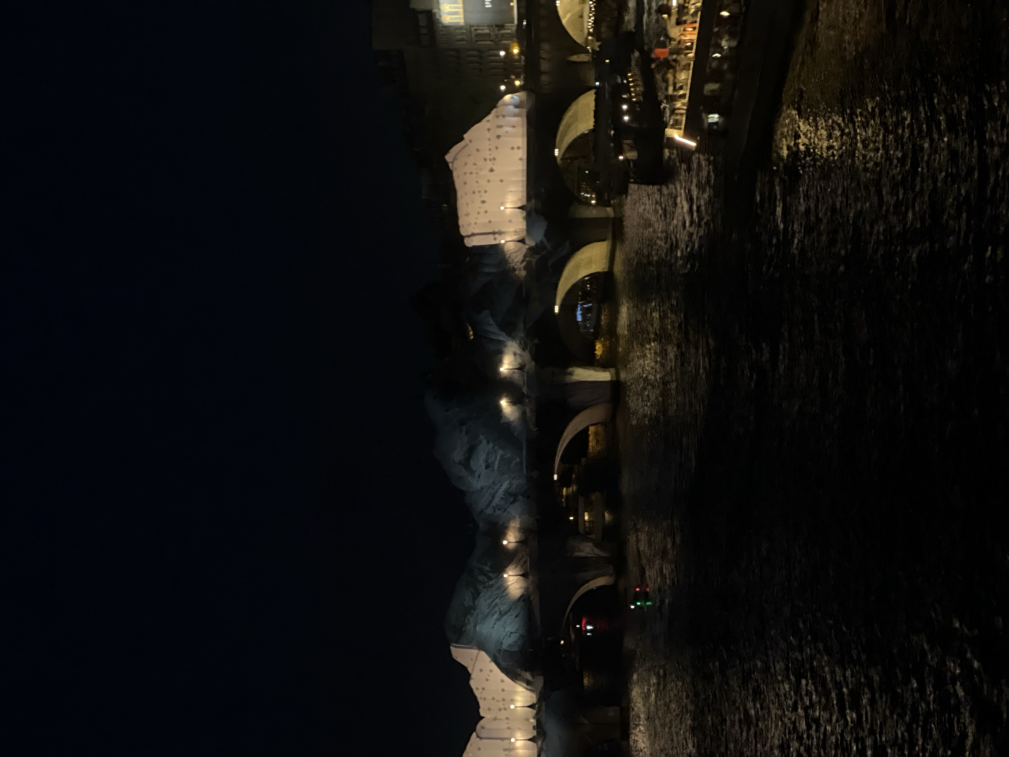 The image captures a nighttime view of a multi-arched stone bridge spanning the River Seine in Paris, France. Prominently featured on or immediately behind the bridge structure are large, illuminated art installations resembling abstract white icebergs or mountains, interspersed with darker, naturalistic mountain-like projections. Numerous streetlights with warm yellow illumination are aligned along the top of the bridge. The dark, rippling surface of the river below reflects these lights. On the right side of the river, at least two large, illuminated tour boats are visible; one shows an individual in an orange garment on its upper deck. A smaller vessel with red and green navigation lights is present in the center-left. The far riverbank on the right features illuminated urban buildings, one displaying a bright commercial sign. The sky above is dark and devoid of visible features.