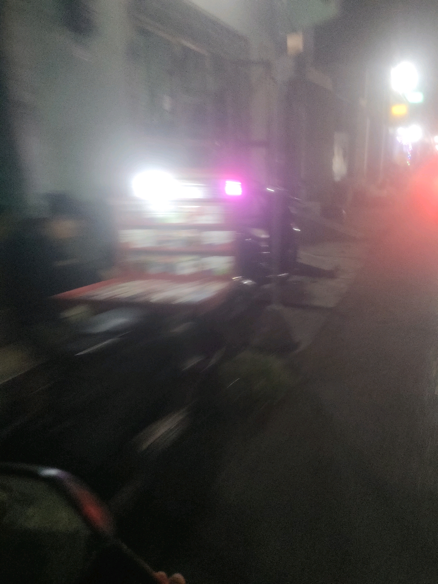 The image is a motion-blurred, nighttime photograph taken from a moving vehicle, likely a motorcycle, on a street in Surabaya, Indonesia. The scene depicts a street with buildings on the left and right. A brightly lit cart or stall is visible in the center, with glowing pink and white lights. The cart appears to be laden with items, possibly food or goods for sale, indicated by the blurry, multi-colored rectangles on its surface. Streetlights and other illuminated signs are present in the background. The foreground shows a portion of the vehicle from which the photo is taken, including a handlebar and what appears to be a gloved hand. The overall impression is of a busy, vibrant street scene at night in an urban environment.