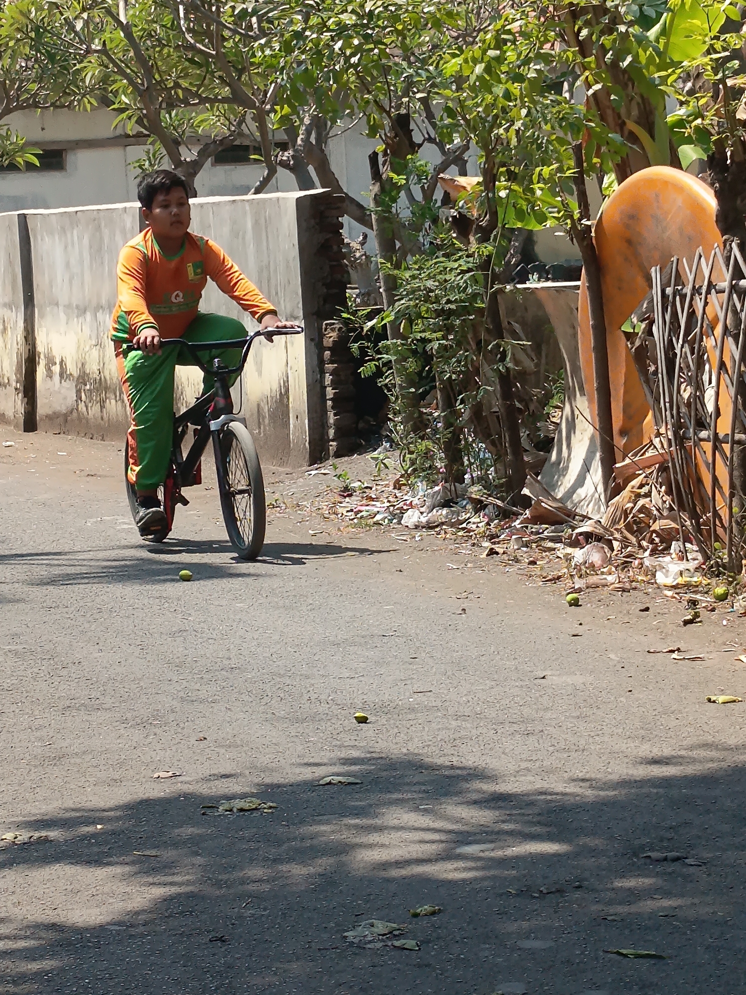 A young boy is riding a bicycle down an asphalt road in Kandangjati Wetan, Indonesia. He is wearing an orange long-sleeved shirt with green accents and green pants. The boy is looking forward and appears to be focused on his ride. The road is lined with a concrete wall on the left and overgrown vegetation and a pile of debris on the right. Sunlight casts shadows on the road, indicating it is a bright day. Several small green objects, possibly fruits or balls, are scattered on the road surface. The background shows buildings and trees, typical of a residential area in the region. The overall scene depicts a casual outdoor activity in a somewhat unkempt urban environment.