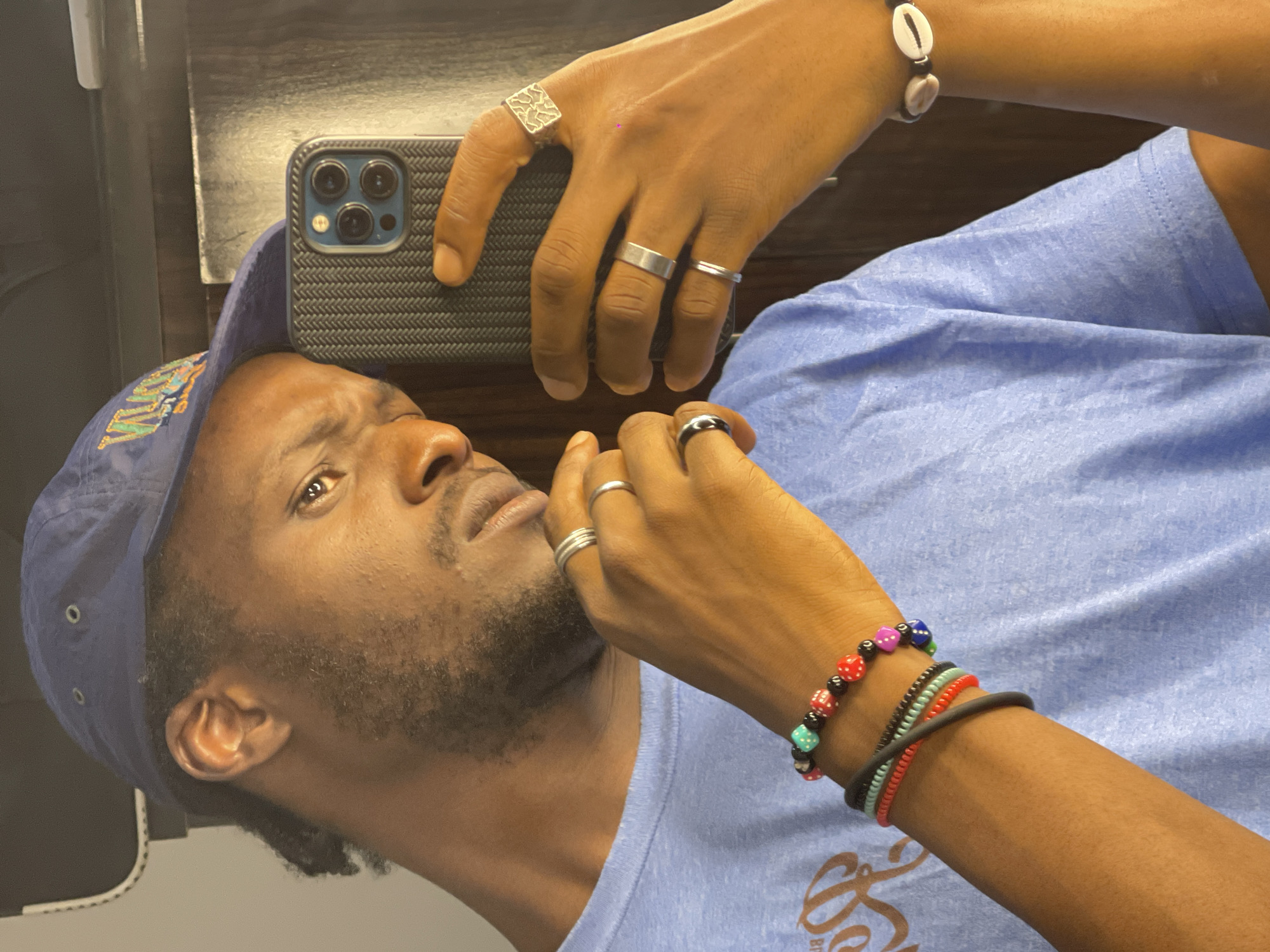 A man is taking a mirror selfie in Abuja, Nigeria. He is wearing a blue baseball cap and a light blue t-shirt. He has several rings on his fingers and bracelets on his wrist. He is holding a smartphone in his right hand, which is also visible in the frame. The background appears to be an indoor setting, possibly a room with a mirror.