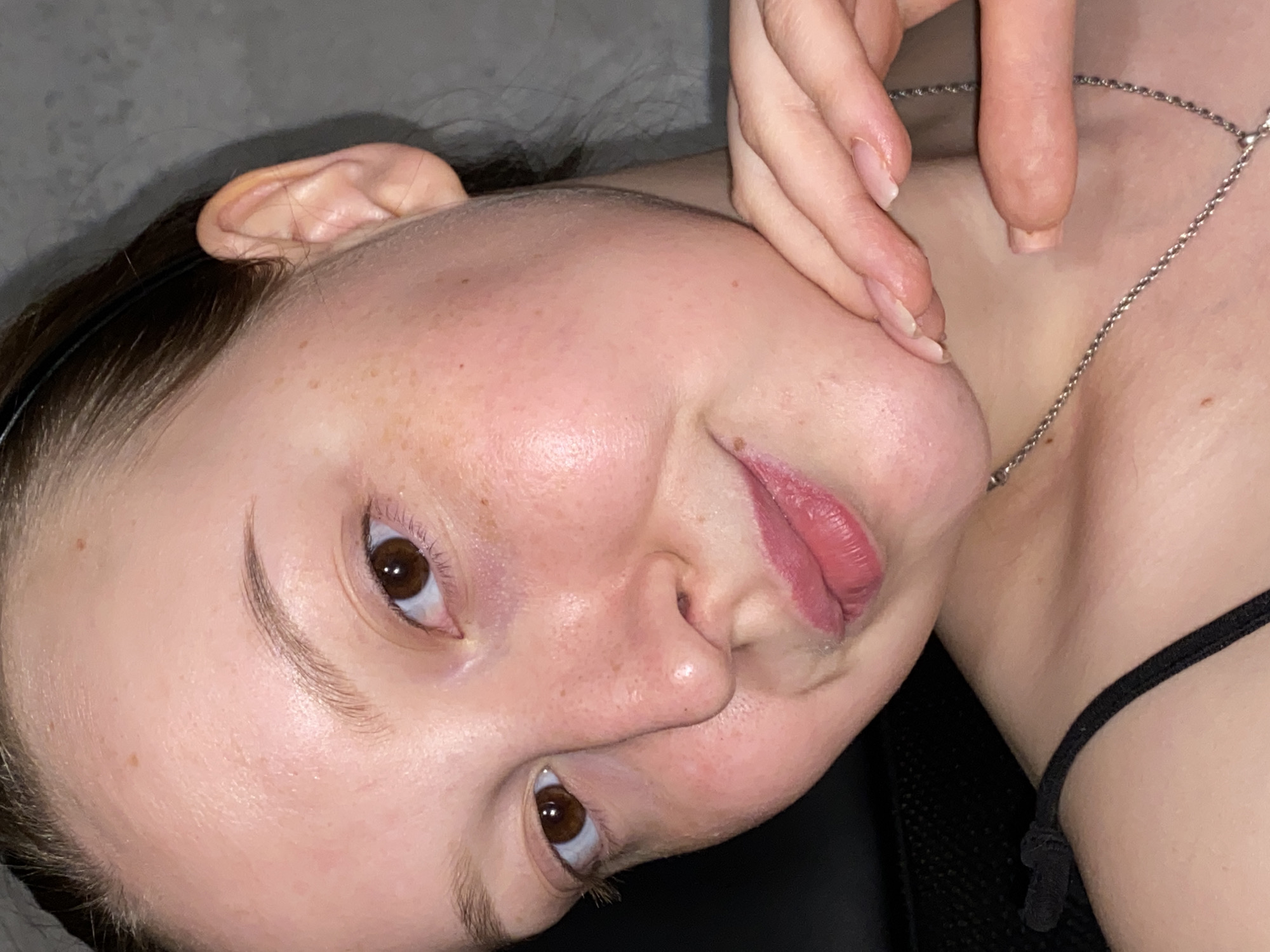 A close-up, slightly angled portrait of a woman with light skin and brown hair pulled back with a black headband. Her facial features include brown eyes, freckles, and pink lips. Her hand is visible, touching her chin. A silver necklace is around her neck, and a black strap of clothing is visible near her shoulder. The background appears to be a gray wall. There is no verifiable evidence to suggest that the image was taken in Chaikovsky, Russia.