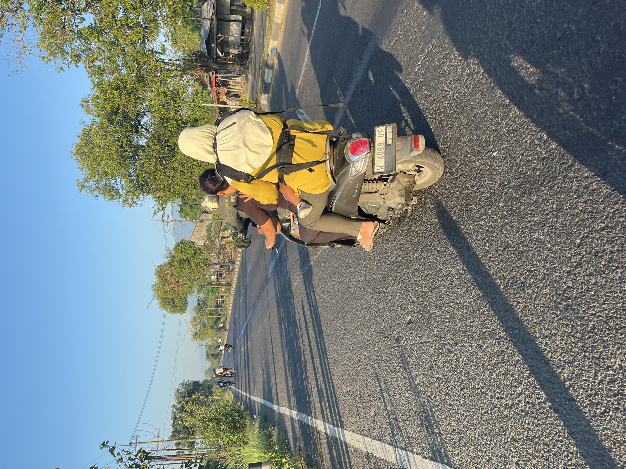 In Ketapang, Indonesia, on a paved road under a clear blue sky, two individuals ride a scooter. The person in the front, wearing a dark shirt and pants, is operating the scooter. Behind them, a person in a yellow top and grey pants, wearing a white hijab and a white backpack, is carrying a young child. The child is wearing a white shirt and grey pants. They are traveling on the right side of the road, with shadows from trees and utility poles stretching across the asphalt. In the distance, other people are seen cycling and riding motorcycles. The environment includes vegetation, trees, utility poles with wires, and buildings lining the side of the road. The scooter's license plate reads "W 4088 UE".