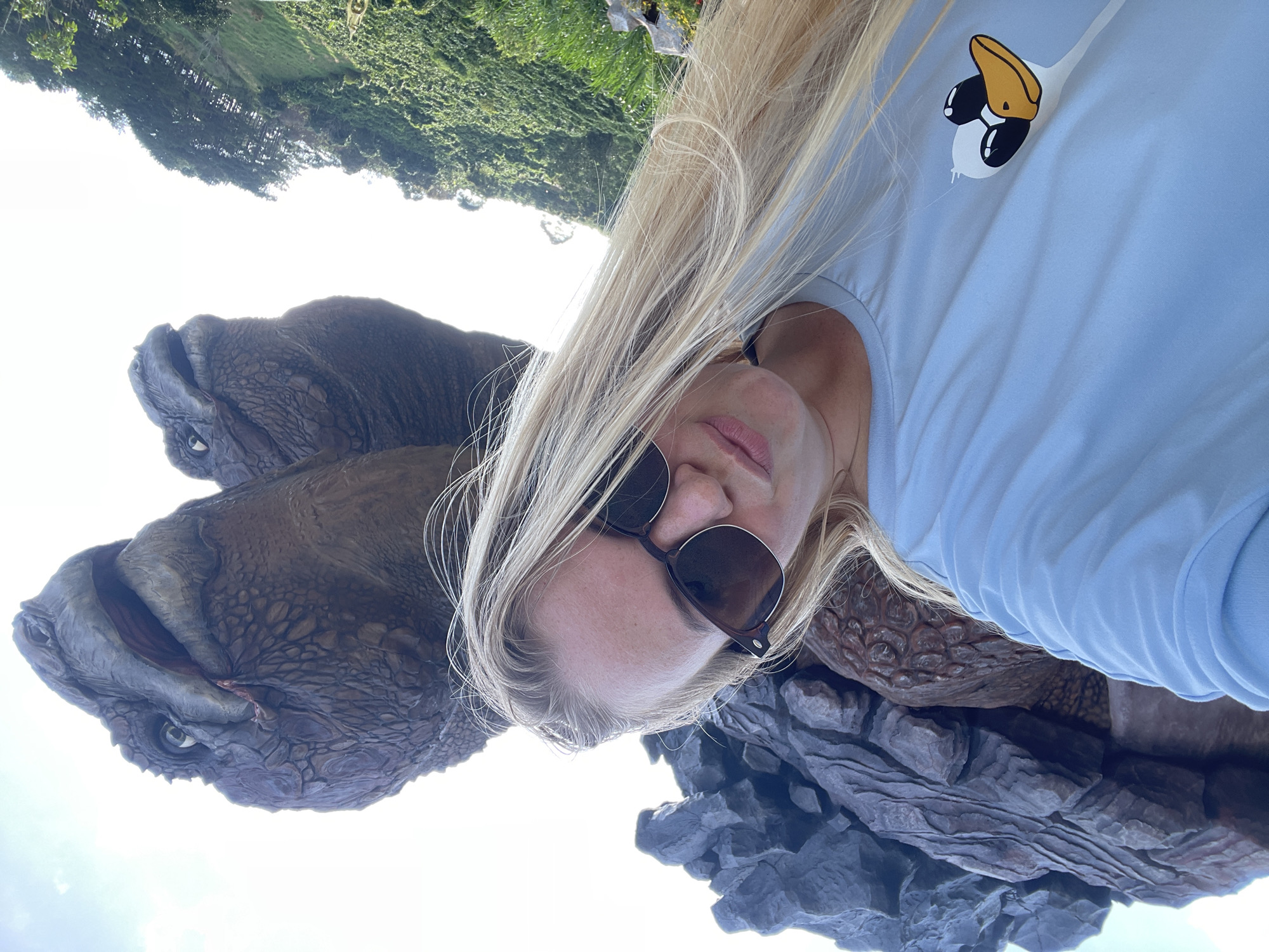In the city of Thôn 1, Vietnam, a young Caucasian woman with long blonde hair and sunglasses takes a selfie. She is wearing a light blue t-shirt with a cartoon duck graphic on the left breast. Behind her, a large, textured rock formation is visible, and two large, dinosaur-like figures with scaly skin and open mouths are positioned above and behind her. Lush green foliage and trees are present on a hillside to the right of the frame, and the sky is bright and overcast.
