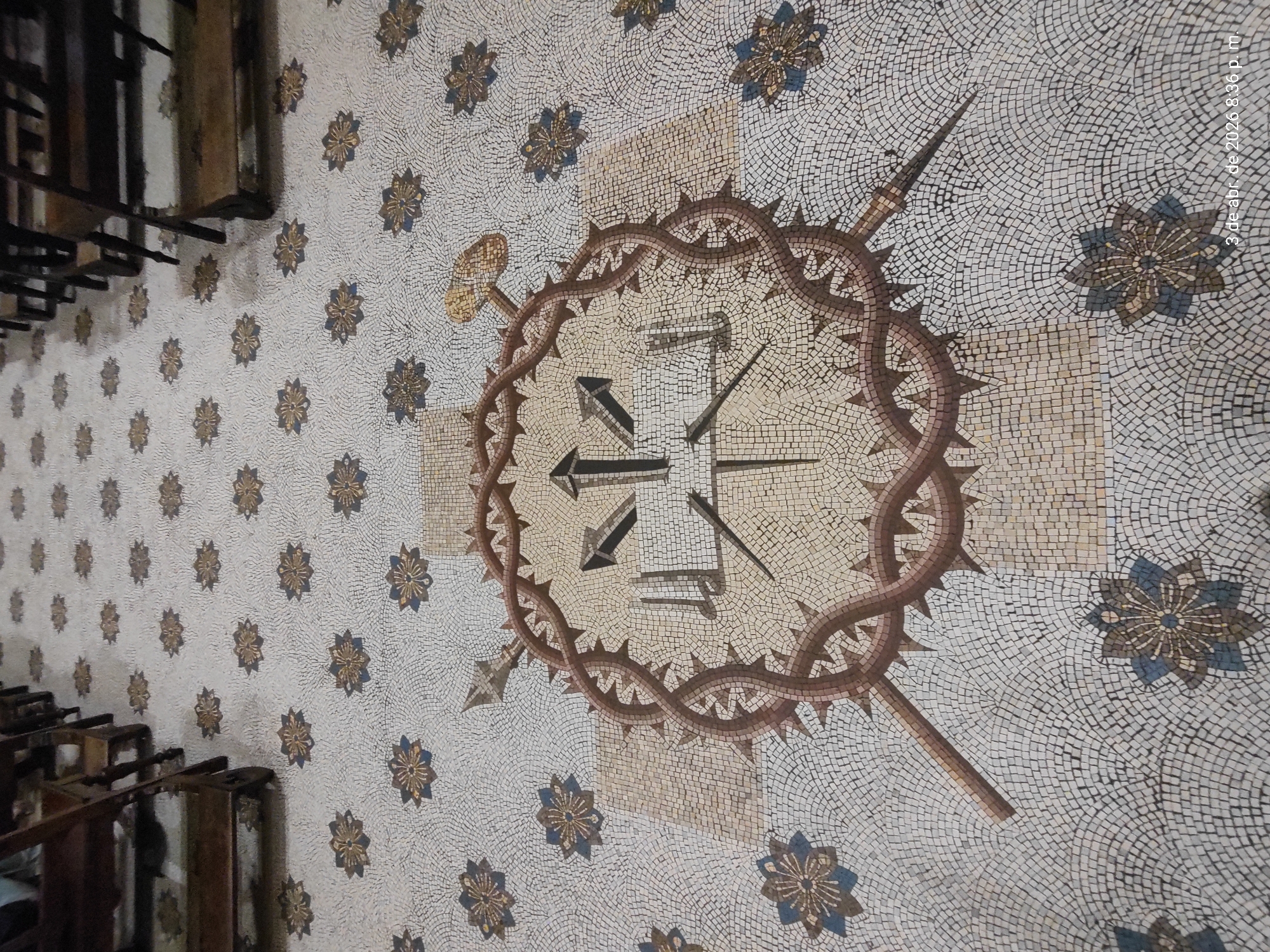 The image shows a mosaic floor in what appears to be a church or religious building. The main design features a representation of the Crown of Thorns, with nails and a spear intersecting it. The background is a field of small, stylized floral patterns in gold and blue on a white base. The floor is likely located in the city of Buenos Aires, Argentina, although the precise location cannot be determined without further information. Seating/pews are partially visible on the left side of the image. There are no people present. The mosaic's color palatte is dominated by neutral tones, brown, gold, and small touches of blue. A date (3 de abr. de 2026 8:36 p. m.) is digitally added onto the image in the upper right.