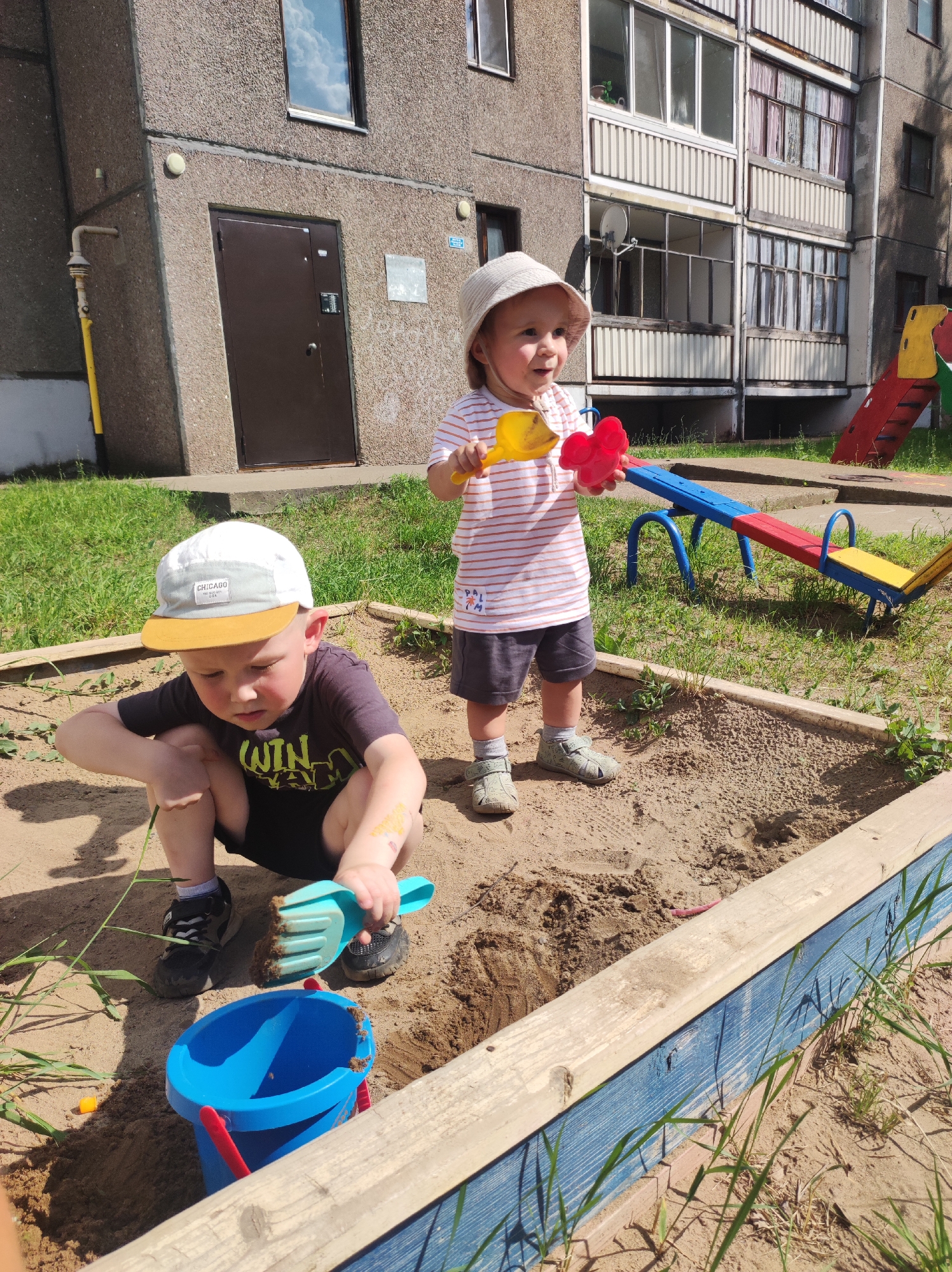 Two young children are playing in a sandbox in front of a multi-story concrete apartment building in Markovsky, Russia. The child on the left, a boy wearing a white and grey baseball cap, a dark t-shirt with a yellow graphic, and black sneakers, is crouching in the sand, holding a blue bucket and a teal shovel. The child on the right, a girl wearing a light-colored sun hat, a striped t-shirt, dark shorts, and grey sandals, is standing and holding yellow and red plastic toys. The sandbox has wooden borders painted blue and brown. In the background, a red, yellow, and blue seesaw is visible on a patch of grass. The building appears to be an older Soviet-era apartment block with balconies. The weather is sunny.