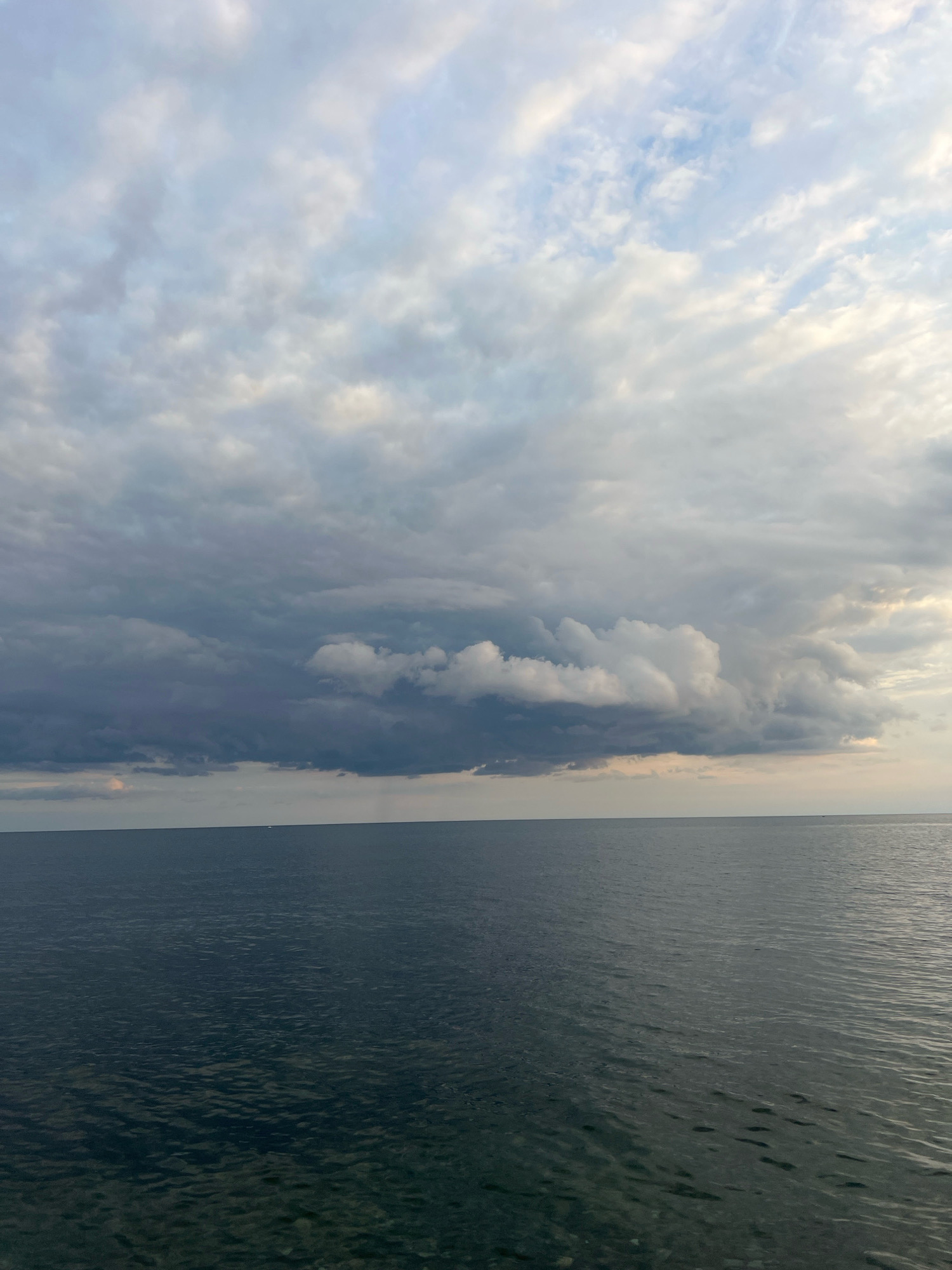 A wide shot captures a vast expanse of dark, rippling water meeting a hazy horizon under a dramatic sky filled with layered clouds. The lower half of the image is dominated by the deep blue-gray water, reflecting the muted light. The upper half showcases the sky, where lighter, wispy clouds intermingle with darker, more ominous formations, suggesting changing weather. A thin line separates the water and sky, with the horizon appearing flat and unbroken. In the far distance, on the horizon, two faint white specks suggest the presence of distant vessels. The overall impression is one of vastness and a potentially turbulent atmosphere.