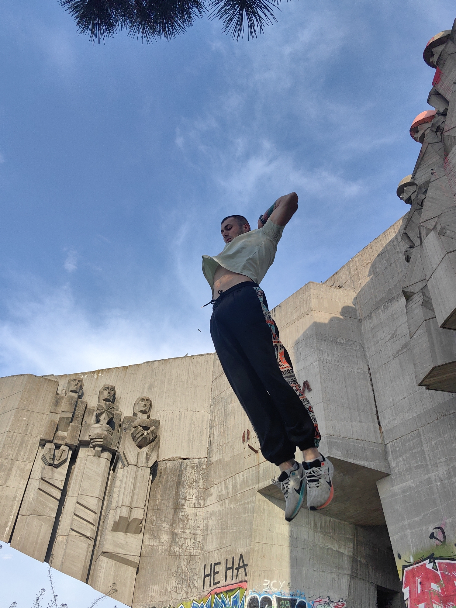 A low-angle shot captures a man in mid-air against a backdrop of a weathered concrete structure, likely a monument or building in Varna, Bulgaria. The man is fair-skinned, bald with a trimmed buzz cut, and appears to be jumping or performing an athletic maneuver. He wears a short-sleeved, light green shirt that reveals his midriff, black pants with a patterned design along the sides, and gray athletic shoes with colored soles. His arms are bent with hands near his head. The concrete structure behind him features bas-relief carvings of human figures and is marked with graffiti. The sky is blue with scattered clouds, and tree branches are visible at the very top of the frame.