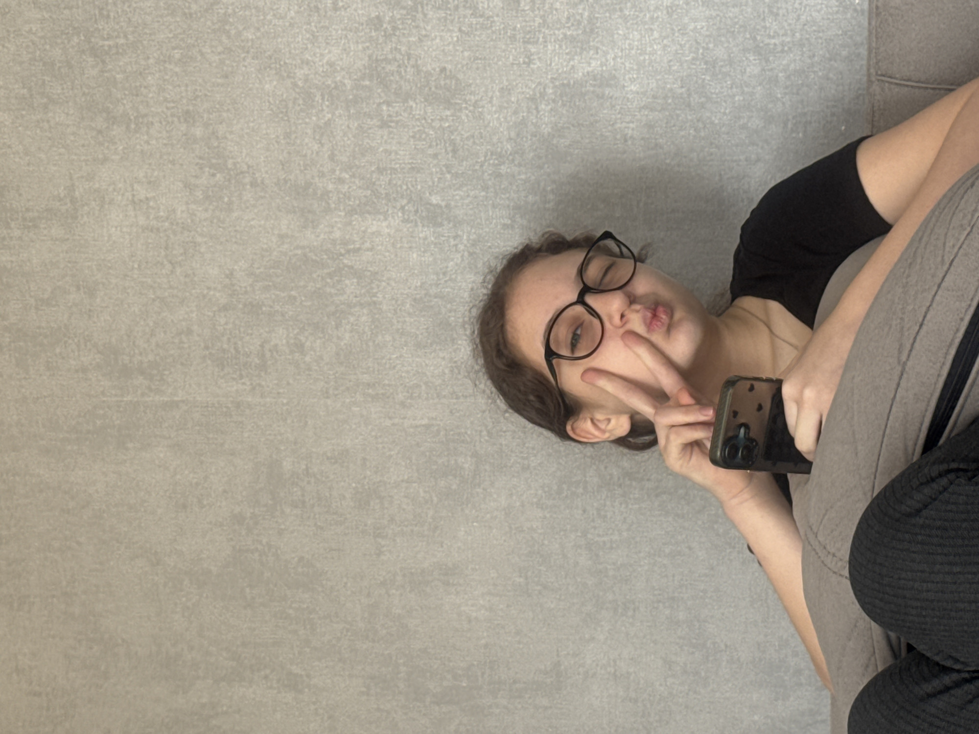 A young woman wearing black-rimmed glasses is positioned on the right side of the frame, making a peace sign with her left hand near her face and pursing her lips. She is holding a black smartphone. She is wearing a black t-shirt and is reclining on what appears to be a gray sofa. A textured gray wall occupies the majority of the left side of the frame. The location is undetermined, but the prompt suggests Tbilisi, Georgia.