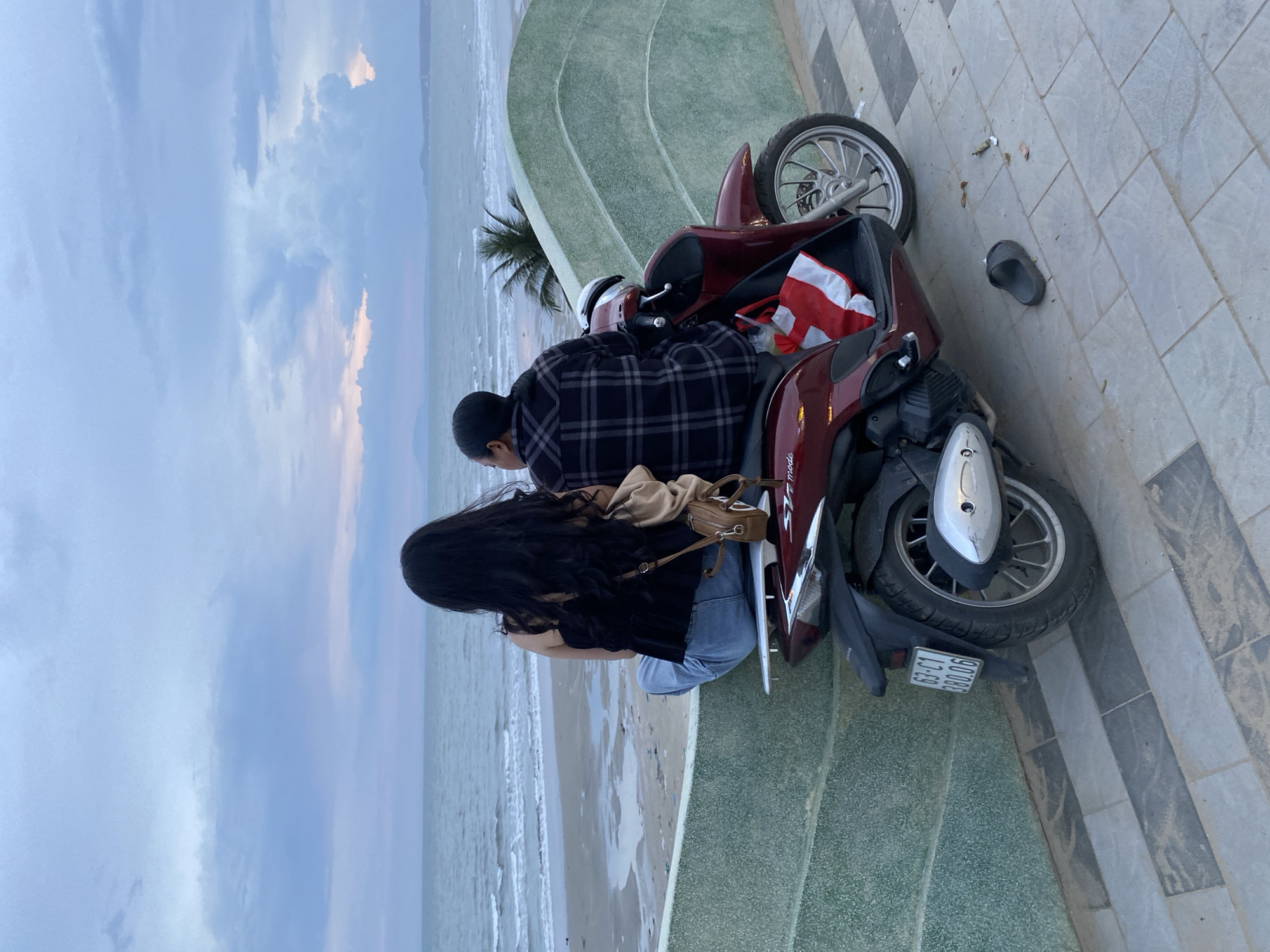 In Mũi Né, Vietnam, two individuals are seated on a red scooter, positioned facing the ocean. The person on the left, a woman with long dark hair, wears a black sleeveless top and blue jeans, and has a tan backpack slung over her shoulder. The person on the right, a man, is dressed in a plaid shirt. The scooter is parked on a paved area next to a textured green wall that curves towards the beach. The sea is visible in the background with gentle waves and a sandy shore. The sky is overcast with hints of pink and orange from a sunset or sunrise. A red and white bag is stored on the scooter.