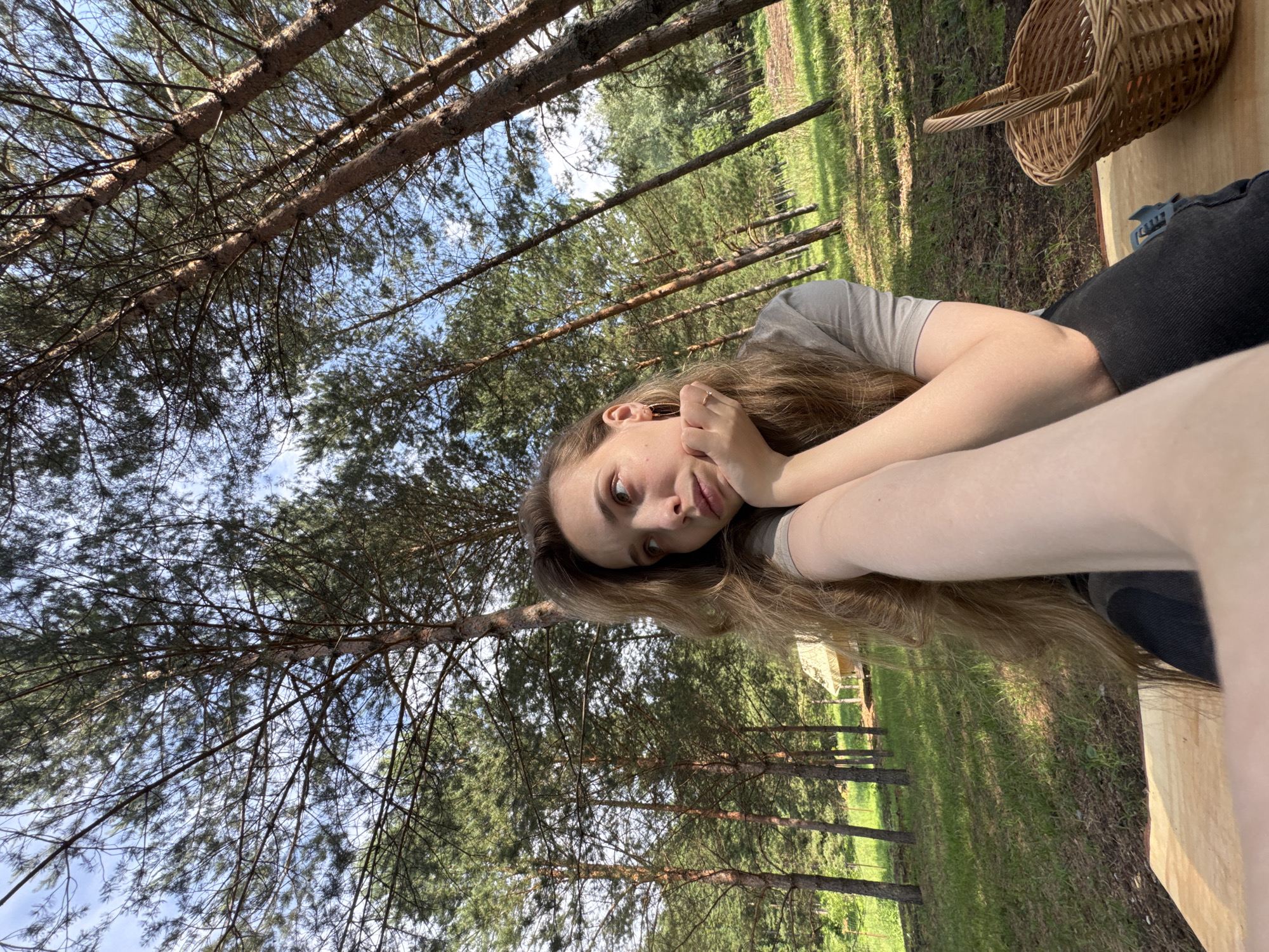A young woman with long, light brown hair is lying down outdoors, with her head propped on her hands. She is wearing a light gray t-shirt and dark pants. Her expression is relaxed and contemplative. Behind her, a woven basket sits on a wooden surface. The setting is a wooded area with tall pine trees and a grassy field. The sun is shining, creating dappled light and shadows on the ground. The scene appears to be taking place on a clear day. The overall mood is peaceful and serene, suggesting a moment of quiet enjoyment in nature. The location context provided is "Гольяны, Russia". There is no visible text in the image.