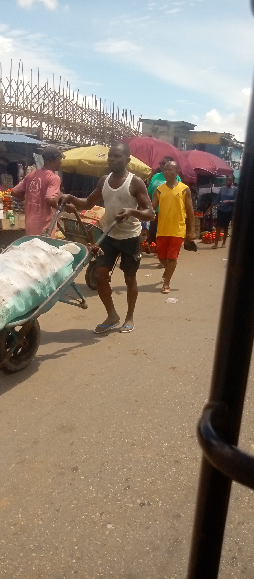 The photograph shows a street scene at a market in an unidentified city. The main subject is a man wearing a white tank top and black shorts, pushing a wheelbarrow filled with a large bag covered in white material. There are other people walking around the market. Another man in a yellow tank top and red shorts is walking to the right of the man with the wheelbarrow. A person wearing a red shirt is to the left of the wheelbarrow. Overhead, there are various market structures, including canopies and wooden scaffolding. The street surface is gray and weathered. The photograph appears to have been taken from inside a vehicle, with part of the vehicle's interior visible in the lower right corner.