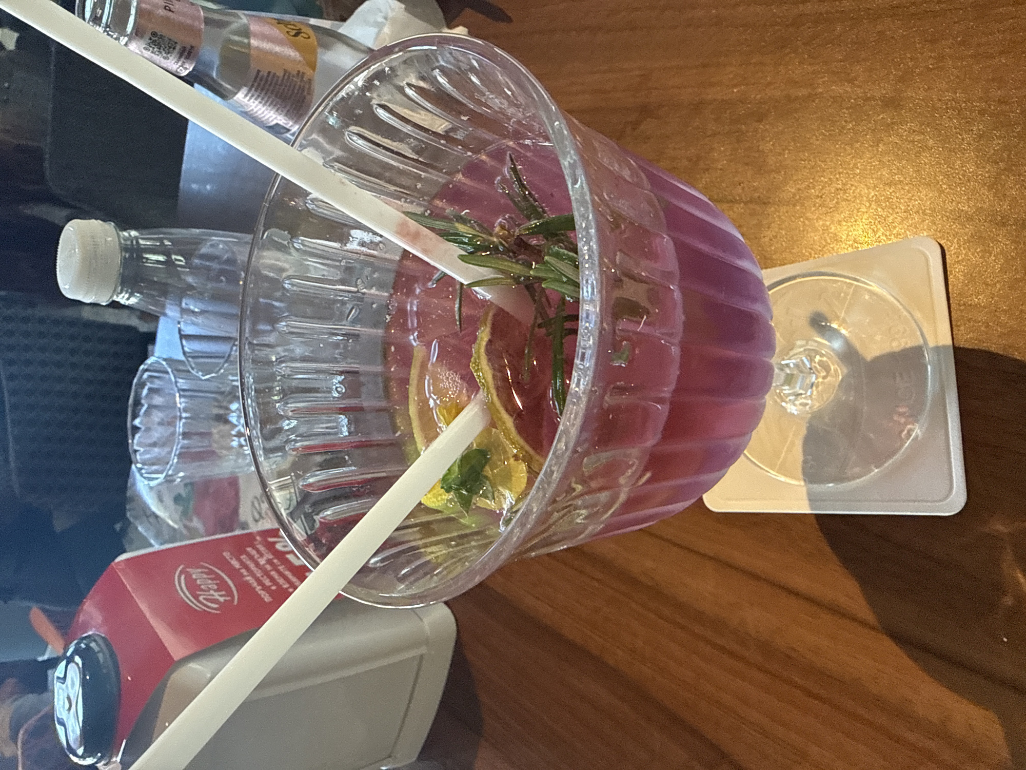 A close-up shot shows a festive purple cocktail in a wide-rimmed glass with two white straws, garnished with slices of citrus fruit and rosemary sprigs. The drink sits on a white coaster on a wooden table. In the background, out of focus, are other drinks, a red carton with the word "Happy" visible, and a clear glass bottle. The scene appears to be at a cafe or restaurant in Plovdiv, Bulgaria, possibly at a venue with a relaxed atmosphere.