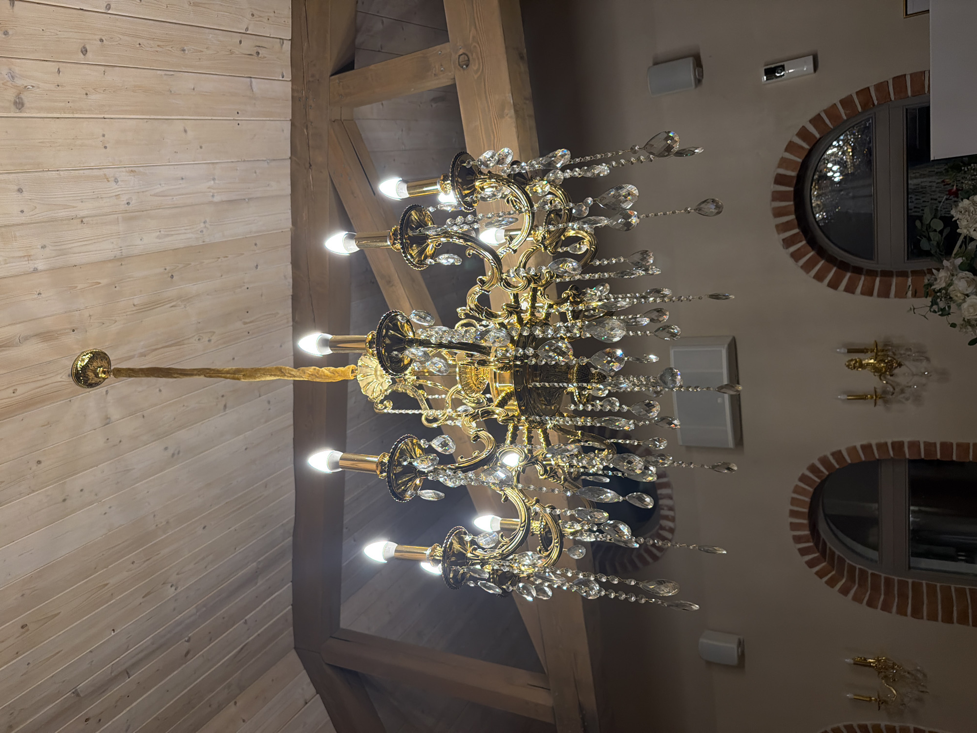 This is a medium shot, eye-level, indoor photograph of a grand, ornate, gold-colored chandelier with numerous crystal accents hanging from a wooden planked ceiling. The chandelier has multiple arms, each holding a light bulb that is lit, and is adorned with teardrop-shaped crystals that refract light. The ceiling features exposed wooden beams, adding to the rustic yet elegant ambiance. Below the chandelier, the wall is painted a neutral tone and features two arched windows with brick trim. Several wall-mounted speakers and what appears to be a security camera are visible on the wall. In the foreground, part of a floral arrangement is visible. The overall setting suggests an interior space such as a restaurant, banquet hall, or a well-decorated home, possibly in Kocherinovo, Bulgaria. There are no people visible in the image.