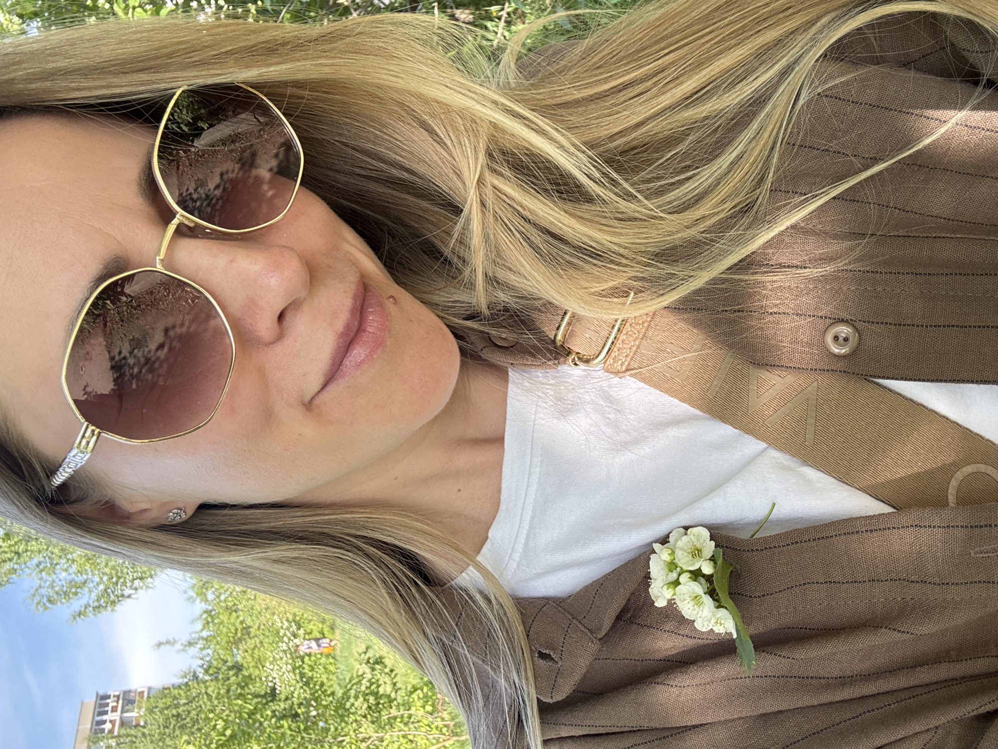 The image shows a woman with blonde hair wearing sunglasses and a brown jacket with a white shirt underneath. A small bouquet of white flowers is pinned to her jacket. The setting appears to be outdoors in a park or green space, possibly in Chaikovsky, Russia, though this is inferred without definitive visual evidence. Details visible in the sunglasses reflect the surrounding outdoor environment, including greenery and structures.