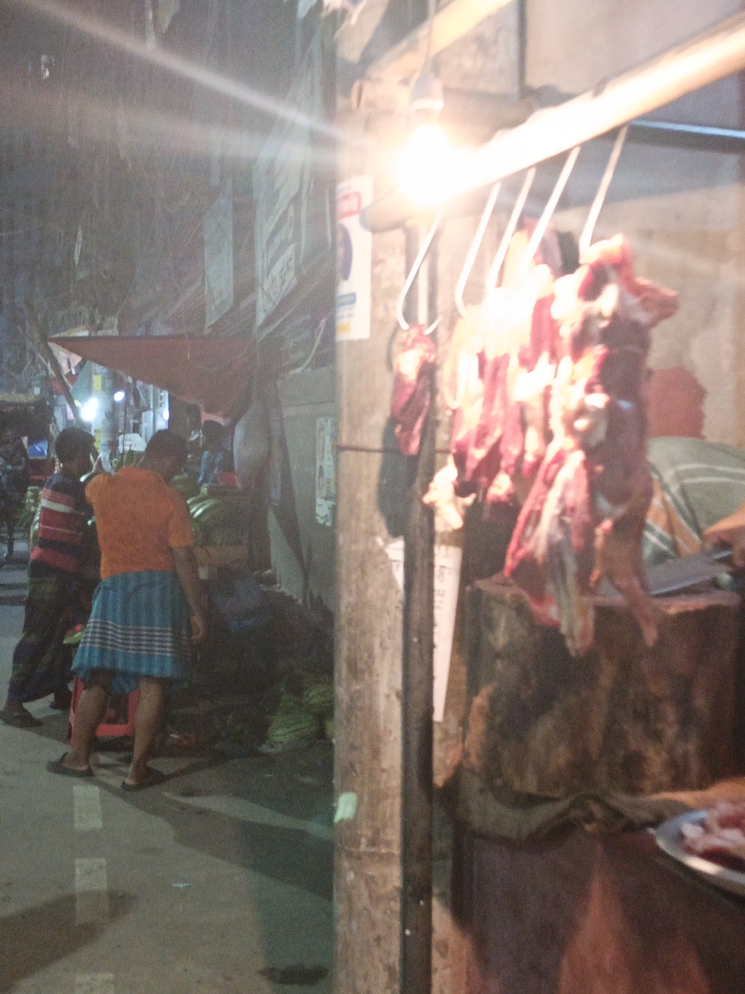 The image captures a vibrant night market scene, likely in Dhaka, Bangladesh. The foreground is dominated by a brightly lit meat stall on the right, where numerous cuts of red meat hang from hooks under a bare light bulb that casts a strong glare across the frame. A large cleaver rests on a wooden chopping block beneath the hanging meat, and a metal tray with smaller pieces of meat is visible at the bottom right.
To the left, several people are present. A man in an orange shirt and a blue and white plaid lungi or skirt stands centrally, facing away from the camera, seemingly tending to a stall. Another man in a striped shirt is visible further left, also with his back to the viewer. In the mid-ground, under a red tarp canopy, are heaps of green produce, possibly bananas or other fruits, suggesting another vendor's stall. Other indistinct figures are visible further in the background.
The ground is concrete, showing shadows and reflections from the various light sources. The scene is illuminated primarily by artificial lights, indicating it is nighttime. The atmosphere appears bustling, characteristic of a South Asian street market. A concrete pillar stands prominently on the right side, with some papers or posters attached, featuring faint, unreadable text, possibly in Bengali script. The overall impression is one of a busy, nocturnal commercial environment.
