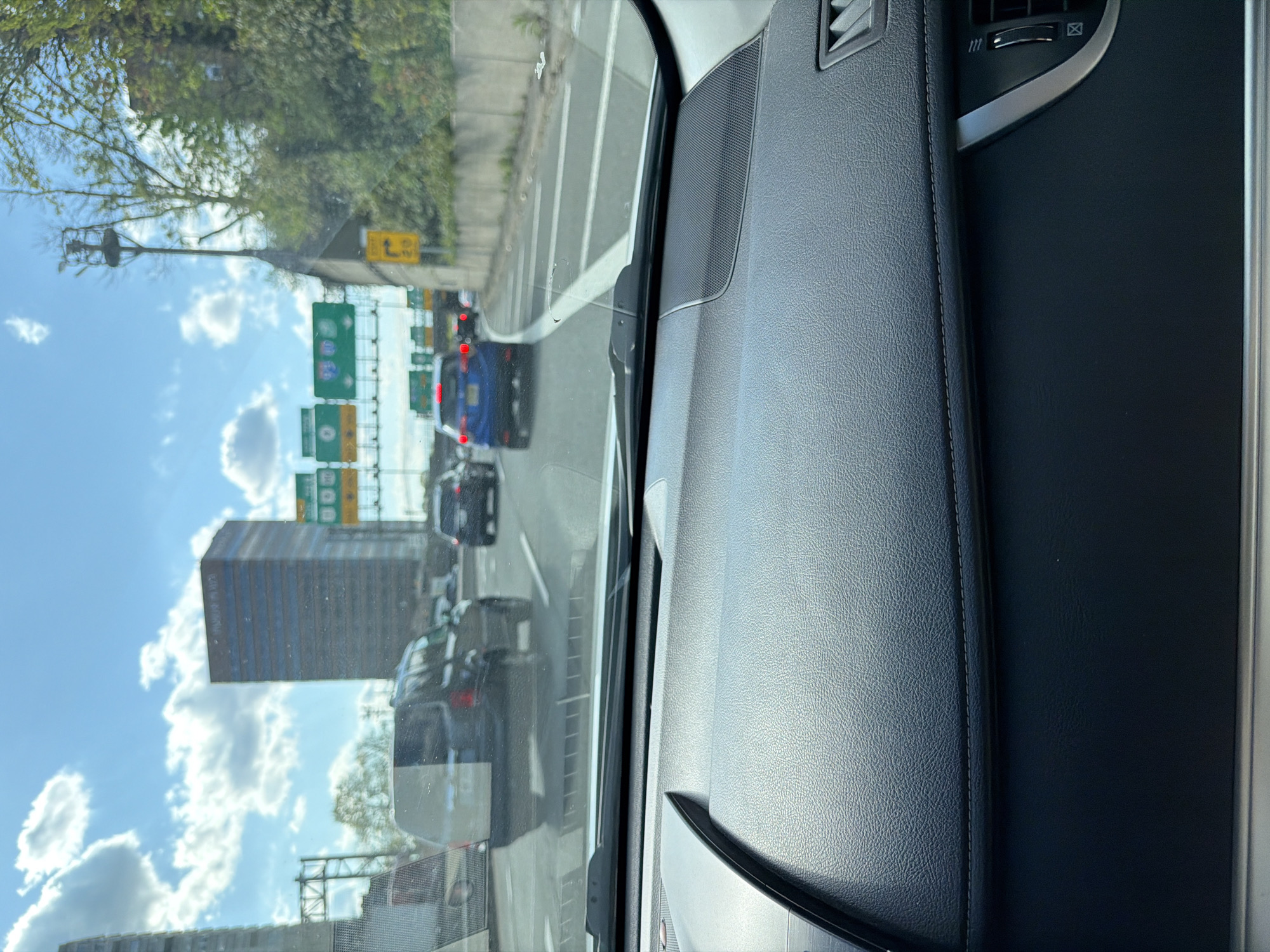 The image, taken from the interior of a vehicle, shows a traffic scene on a highway in Fort Lee, New Jersey, United States of America. The highway appears congested, with multiple vehicles visible, including a blue sedan and a dark-colored van. Signage indicates highway exits and directions. A tall office building is visible in the background. The sky is blue with scattered clouds. The vehicle's dashboard and interior panels are prominently featured in the foreground.