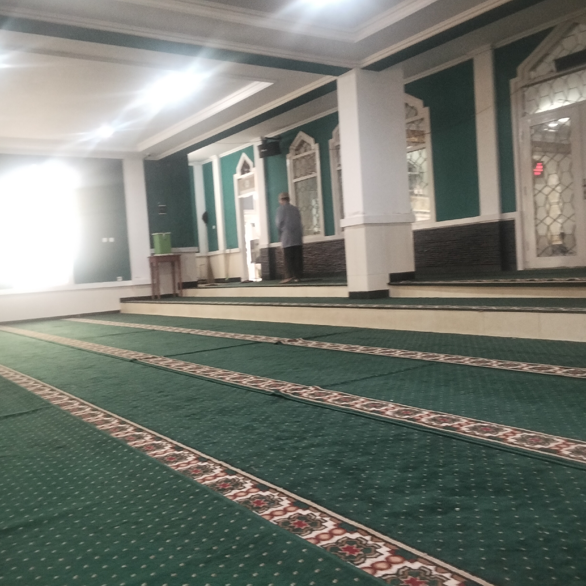 The image shows the interior of a mosque in Cisaat, Indonesia. The main focus is the green carpet with repeating prayer rows indicated by decorative borders. The carpet fills the foreground and extends into the middle ground. The middle ground reveals a raised platform with more carpeted areas and beige borders. A person, seen from behind, stands on this platform facing away from the camera, dressed in a light-colored shirt and dark trousers. To the left of the person, a small wooden table with a green container on it is visible. The background features walls painted in alternating sections of green and white, with decorative white trim. Several arched windows with decorative grilles are present on the walls. White columns support a white ceiling with visible lighting fixtures. The overall impression is a clean and well-maintained prayer space.