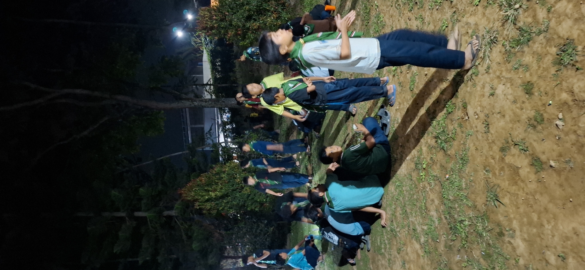 A group of young boys, possibly scouts, are gathered in an outdoor area at night. They are wearing dark blue pants and some have green scarves around their necks. Some are sitting on the ground, while others are standing. The ground is covered in dry grass and dirt. There are trees and bushes in the background, and some artificial lighting is visible, suggesting an urban or park environment in Puntukrejo, Indonesia. The overall atmosphere appears to be casual and social, with the boys engaged in conversation or activity.