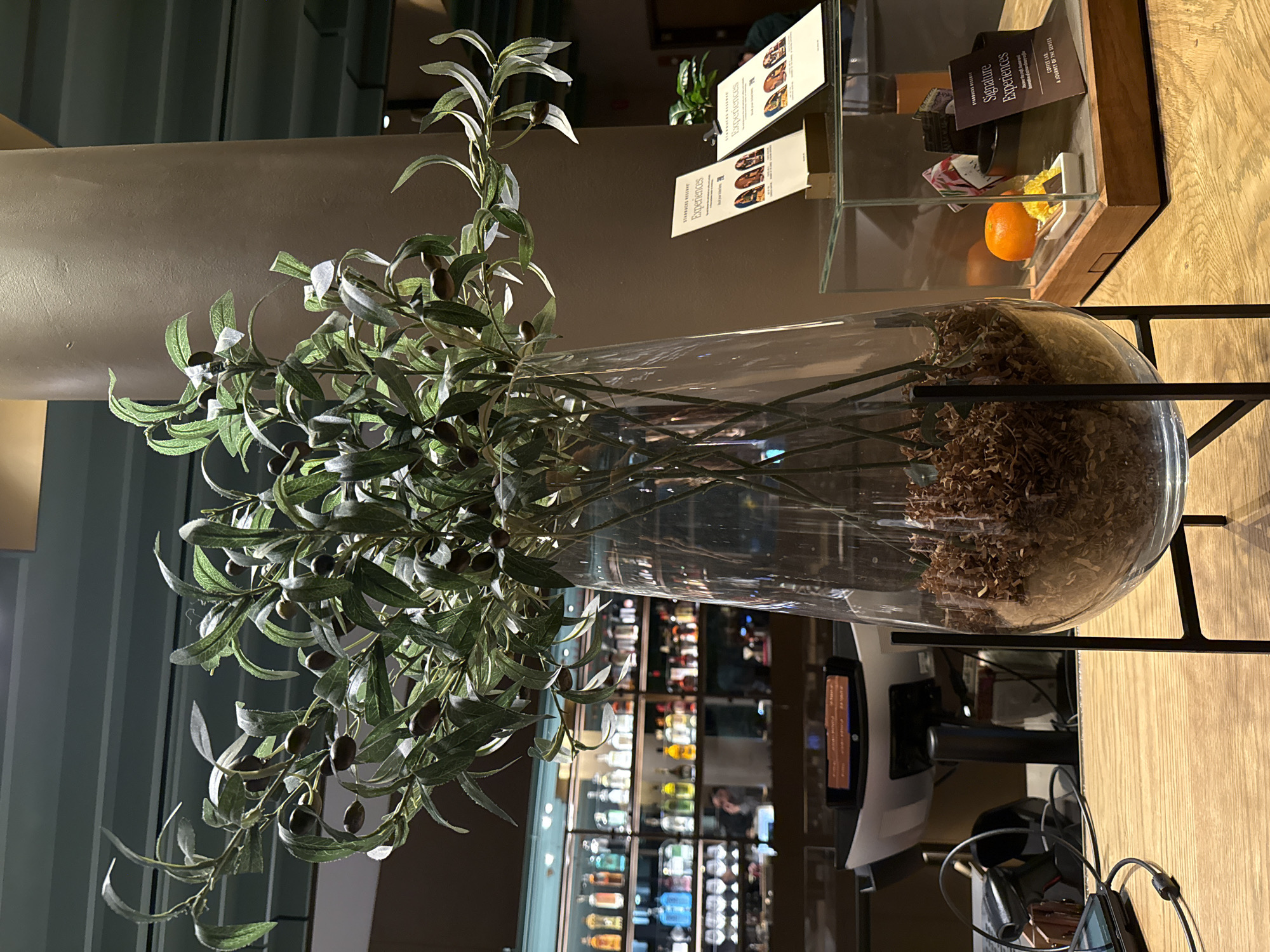 Here is a detailed description of the image:
The photograph depicts an interior scene, potentially of a restaurant or bar, with a focus on decorative elements. In the foreground, there's a large clear glass vase filled with dry, shredded material. A leafy plant with dark olives extends from the vase. The vase rests on a black metal stand.
To the right of the vase is a wooden countertop with a glass display case containing brochures, an orange fruit, and other items. The brochures have the words "STARBUCKS RESERVE Experiences" and "Signature Experiences" printed on them.
In the background, there is a bar area with shelves stocked with bottles. Reflected in the glass of the vase are interior design elements, including a light fixture, and the interior of the bar area.
The location appears to be in a city as suggested by the bar in the background. Judging from the brand, it's likely to be a Starbucks Reserve location. It's difficult to pinpoint the precise location in Chicago without further context or cues from the image itself.
