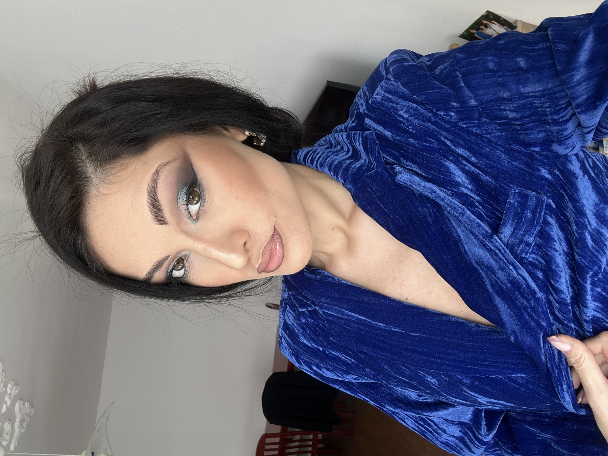 A woman is seen from the chest up, turned slightly to the right, wearing a textured, royal blue robe or jacket. She has dark hair, light skin, and distinctive makeup that includes smoky eyeshadow and carefully shaped eyebrows. Her expression is neutral, and she's looking slightly upward and to the left. The background is nondescript, featuring a light-colored wall and a hint of furniture or objects in the periphery. The location is Unknown, Unknown.