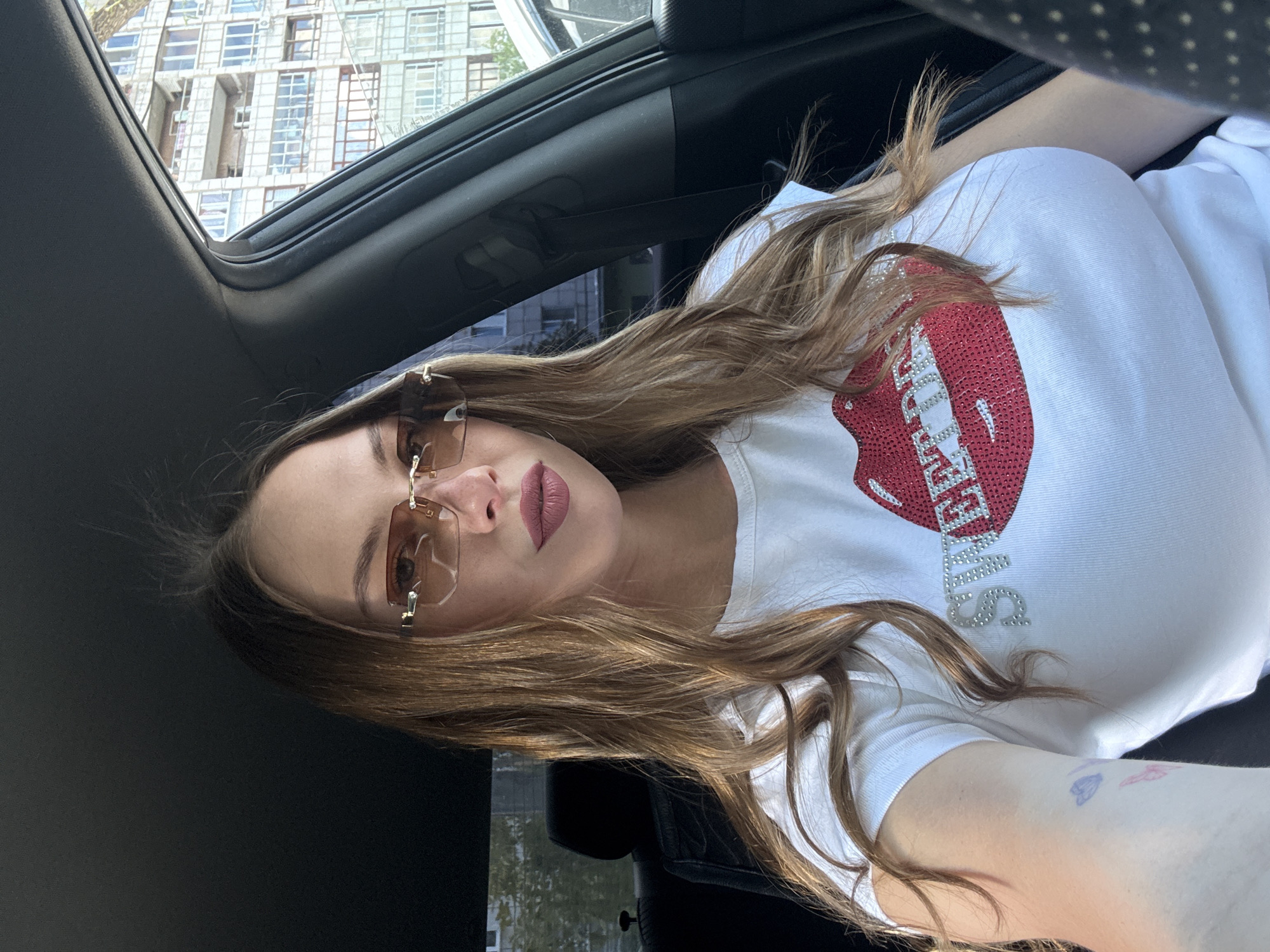 A young woman with long, wavy brown hair and auburn undertones is sitting in the driver's seat of a car. She is wearing rimless, amber-tinted sunglasses and a white t-shirt adorned with a sparkly red sequined image of lips and the word "SWEET" spelled out in silver rhinestones. Her lips are painted with a matte, dusty rose lipstick. A faint, small tattoo of colorful butterflies is visible on her left forearm.
The interior of the car is dark gray, with the black leather of the headrest and seat visible. The window on the right reveals an urban environment. A multi-story apartment building with numerous windows, balconies, and cream-colored facade is prominent. The building's architecture suggests it is from the Soviet era. Trees are visible in front of and to the side of the building. The sky appears bright and sunny. The car appears to be parked on a street or in a parking lot in the city of Perm, Russia, based on the architectural style of the building.