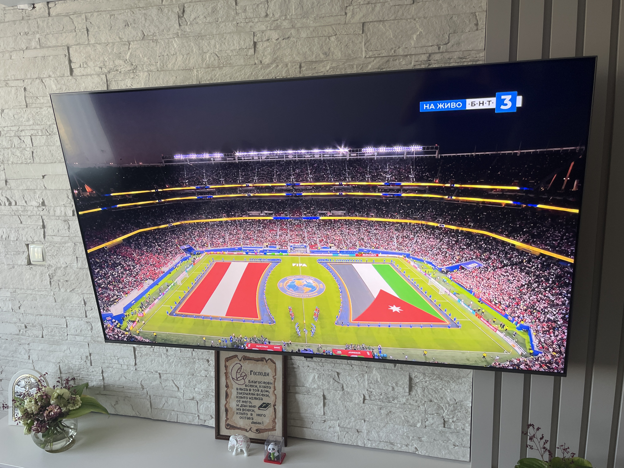 A large, flat-screen television is mounted on a white stone-tiled wall in a room, likely a living room, located in Varna, Bulgaria. The television displays a live broadcast of a soccer match played in a stadium packed with spectators. The field features a large FIFA World Cup Games logo at its center. Two national flags are prominently displayed on the field, one appearing to be Austria's and the other Jordan's. A channel logo with the text "НА ЖИВО" (Live) and "Б.Н.Т. 3" is visible in the top right corner of the screen. Below the television, on a white shelf, are decorative items: a glass vase with a floral arrangement, a framed embroidered text, a small white elephant figurine, and a small panda figurine. The wall to the right of the television has vertical gray paneling.