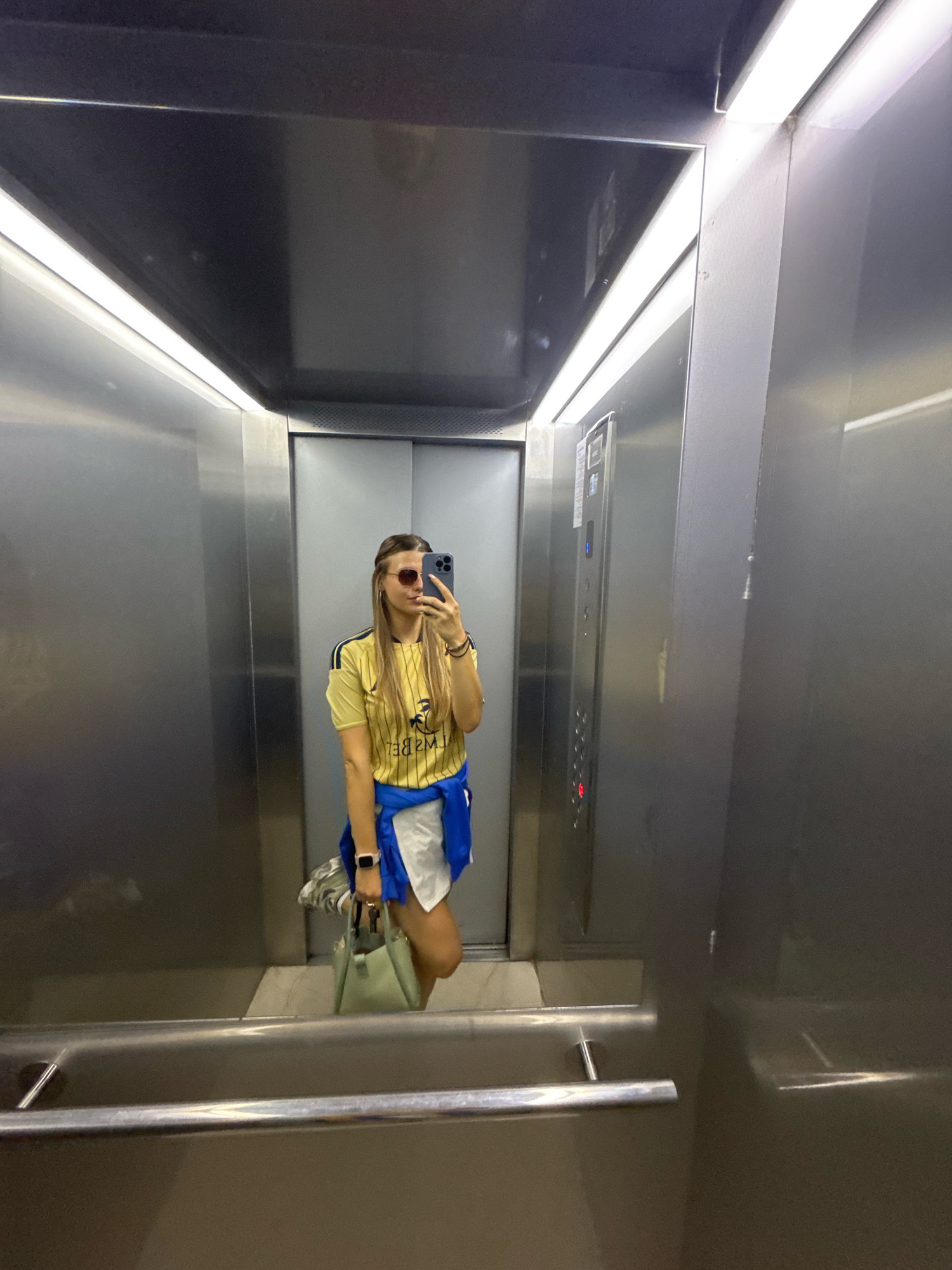 A young woman with long blonde hair pulled back in a ponytail, wearing sunglasses, a yellow striped t-shirt, and a white skirt with a blue jacket tied around her waist, stands in an elevator and takes a selfie with her phone. She is carrying a light green purse. The elevator is made of brushed metal and has bright fluorescent lighting. The elevator control panel is visible on the right wall. The mirror reflects the woman and the elevator interior.
