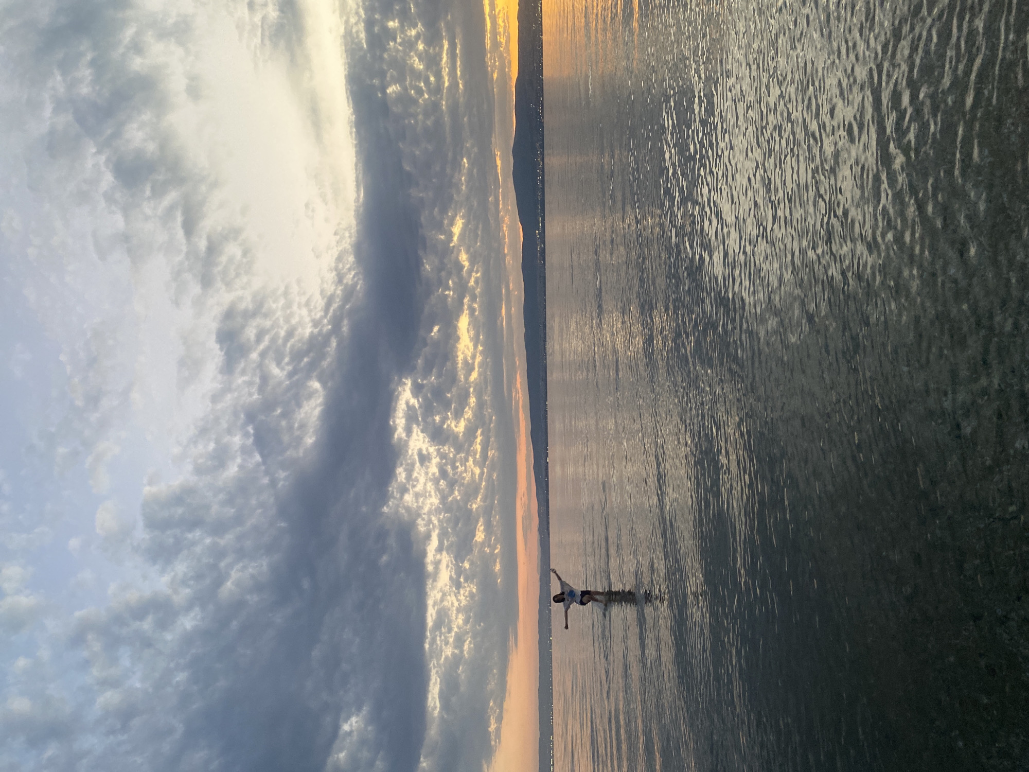 The image depicts a serene sunset over the Thermaic Gulf, as seen from the coast of Neoi Epivates, Greece. A young person, clad in a white t-shirt and dark shorts, stands in the shallow, calm waters, with arms outstretched and legs apart in a dynamic pose. The water reflects the muted colors of the sky, a mix of pale blue, grey, and hints of orange and yellow from the setting sun. In the distance, a hazy silhouette of a coastline with rolling hills and scattered lights marks the horizon. The sky is adorned with a dramatic array of clouds, ranging from dark grey formations to lighter, wispy clouds catching the last rays of sunlight. The overall atmosphere is peaceful and contemplative, capturing a moment of solitary joy at the edge of the sea.