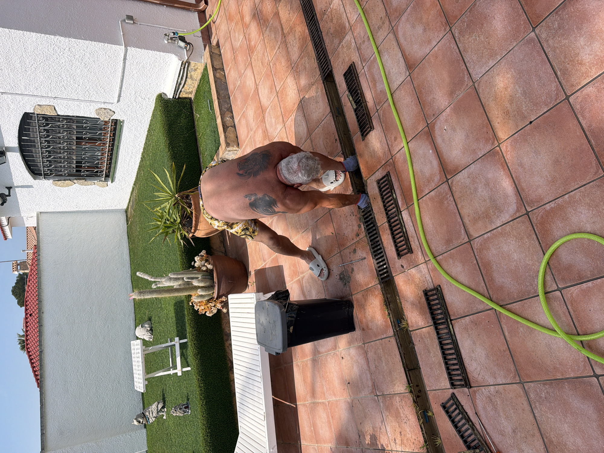 Outside a building in Mont-roig del Camp, Spain, a person is cleaning a drainage channel with a brush. The person has short grey hair and is wearing beach shorts and socks with open toed shoes. The back of the person has tattoos on the left and right shoulder blade. The person is cleaning the channel beside a brick patio. A green water hose stretches across the patio. Behind the patio is a garden with an outdoor bench and potted plants.