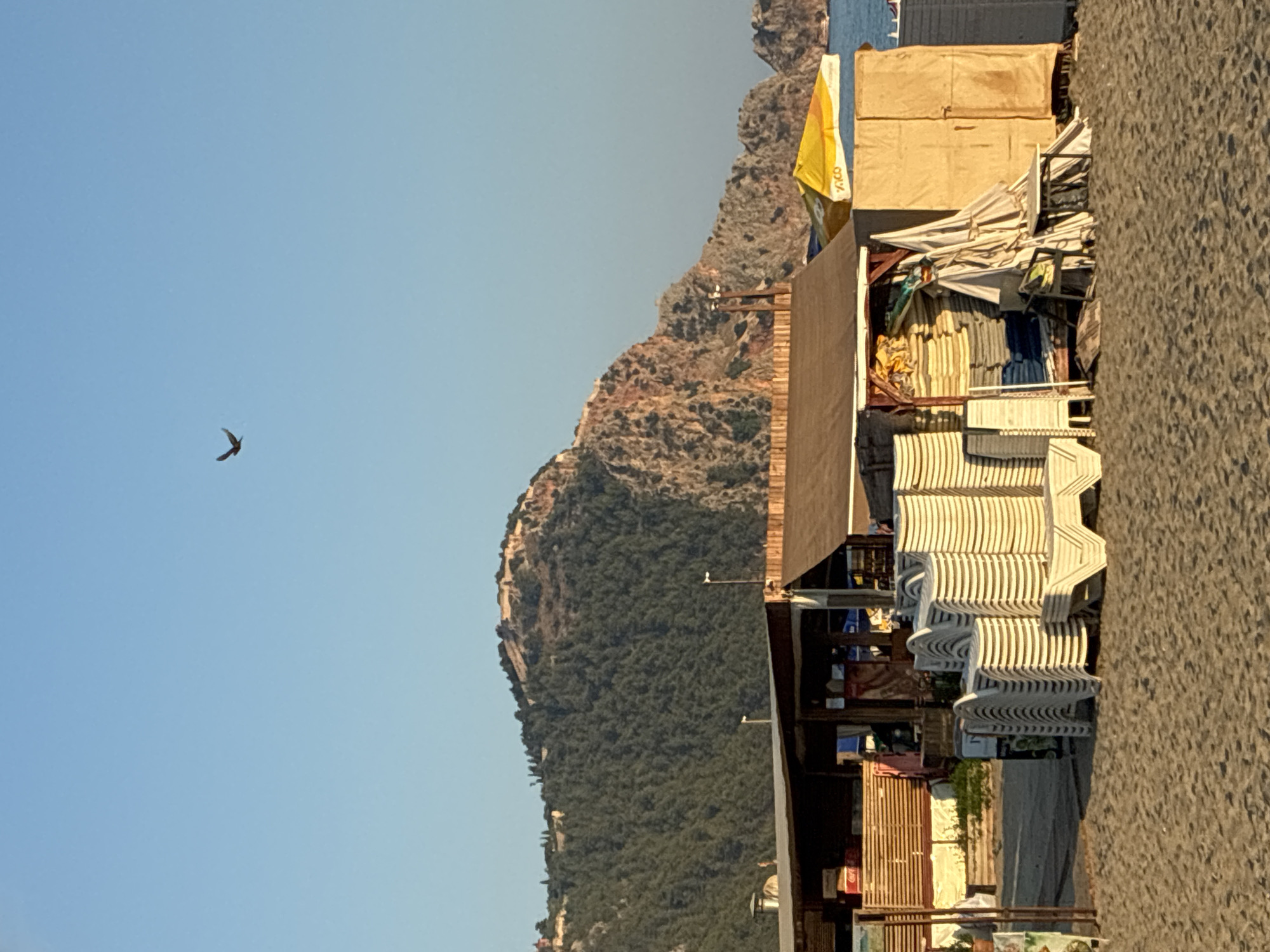 The image depicts a beach scene in Alanya, Turkey, with a cluster of beach chairs and umbrellas stored near a wooden structure. The background features a lush green mountain with ancient fortifications at its summit, characteristic of the Alanya Castle. The sea is visible in the distance, with a boat on the horizon. The sky is clear and blue, with a bird in flight. The foreground consists of sand, suggesting a typical beach environment. The overall impression is one of a serene and picturesque location, likely during daylight hours.