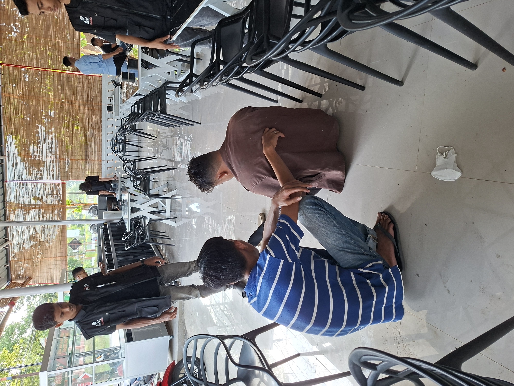 The image shows an indoor area in Lhokseumawe, Indonesia. There are multiple people present. Two men are on the floor, appearing to be engaging in some kind of interaction. One is wearing a brown shirt and the other a blue and white striped shirt. Several men are standing, wearing dark shirts with logos or designs, possibly uniforms. Stacks of black plastic chairs are visible. A discarded face mask sits on the tiled floor. Through windows, greenery and buildings are seen.